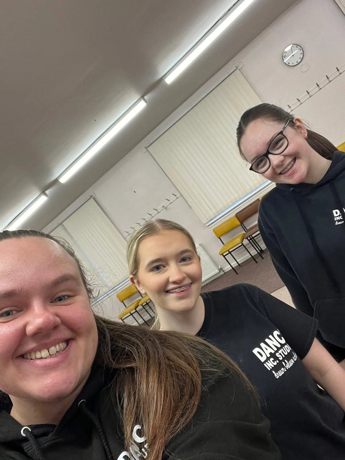 Three young girls smiling for a selfie in a classroom, with chairs stacked against the wall and a clock showing 2:16.