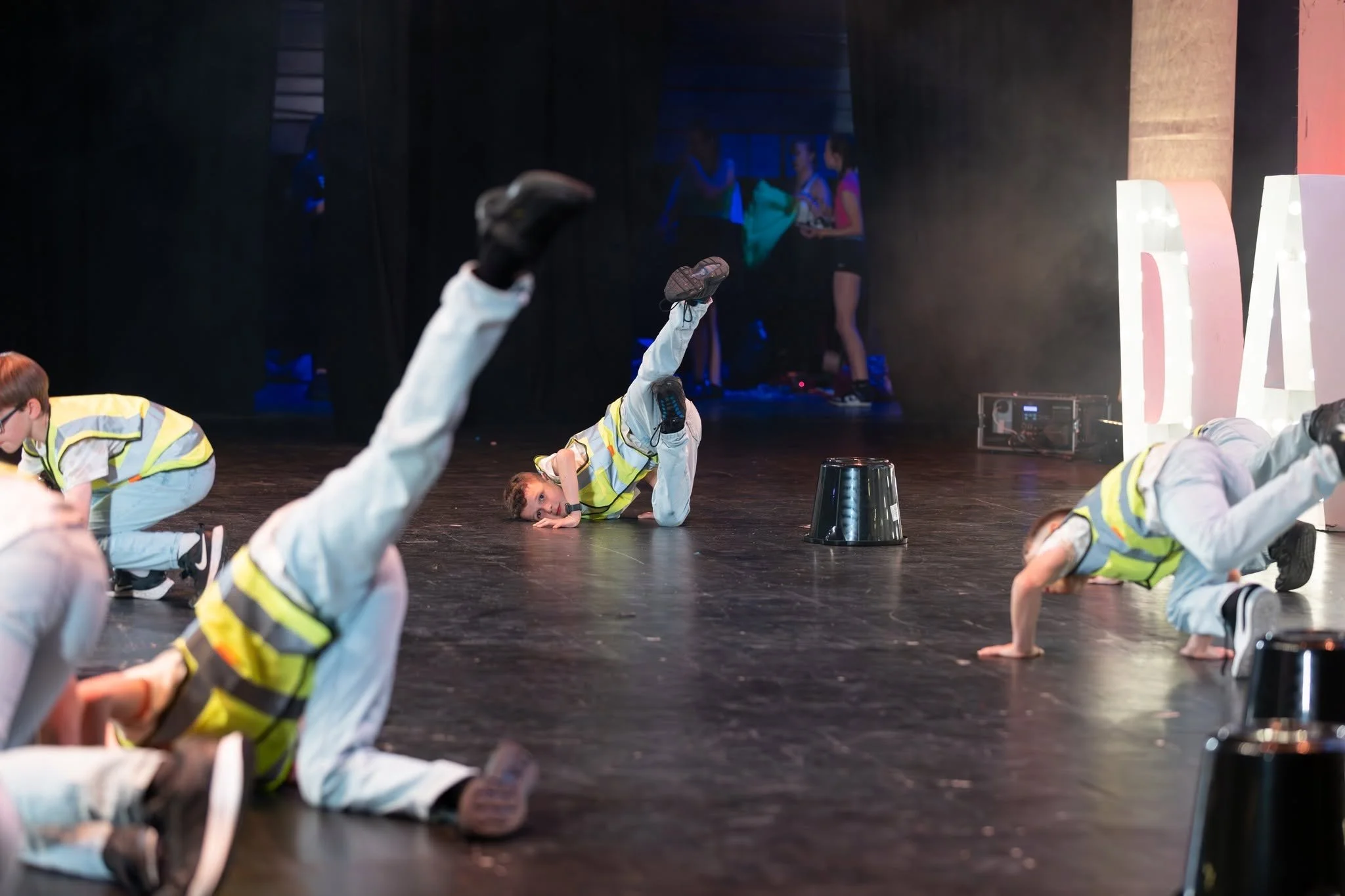 Children performing breakdance on stage, wearing yellow safety vests and white pants, with a dark background and a group of young women in casual clothes standing in the background.