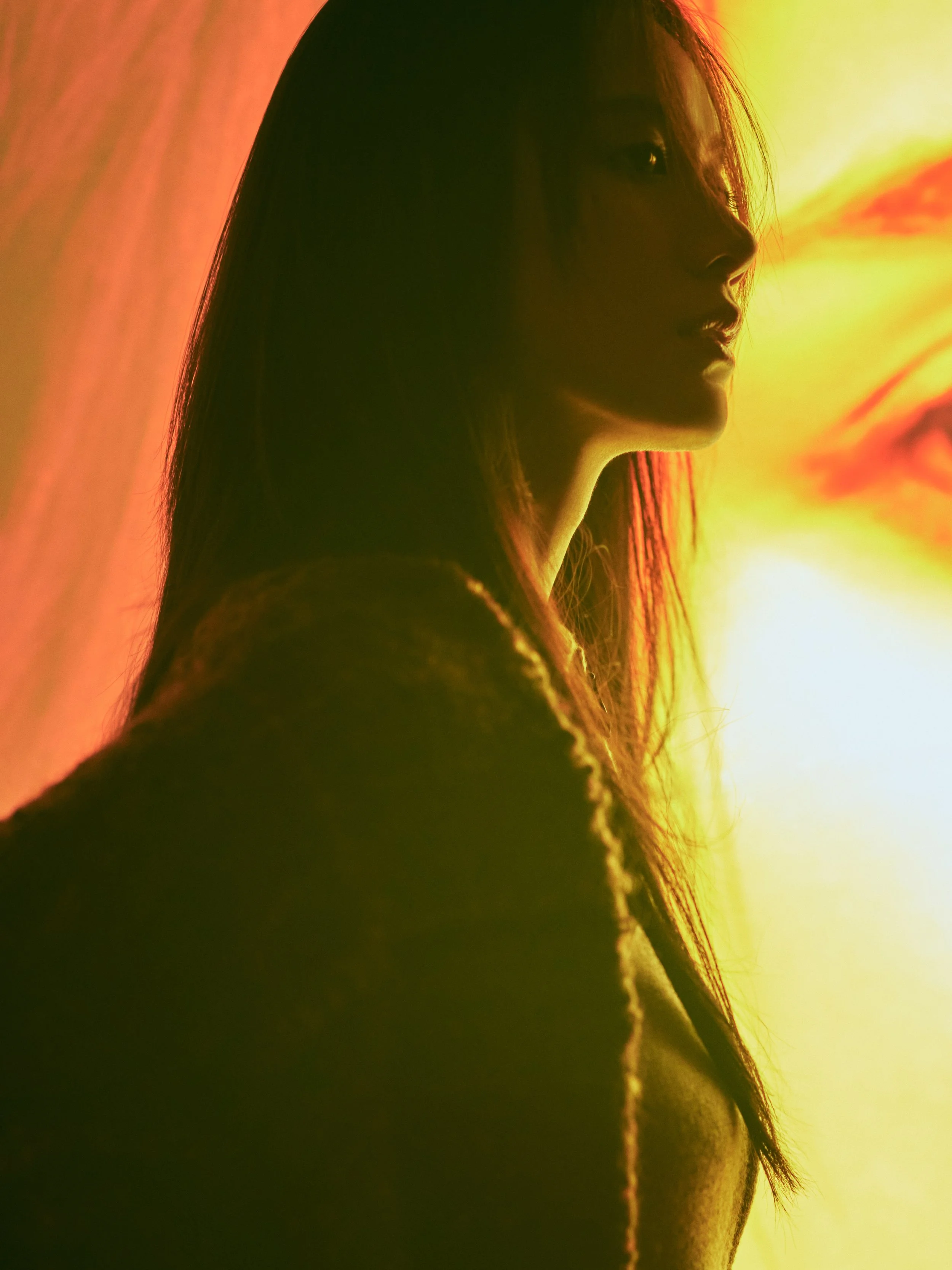 A silhouette of a woman's face in profile, illuminated by warm yellow and orange lighting, with long hair visible against a colorful background.