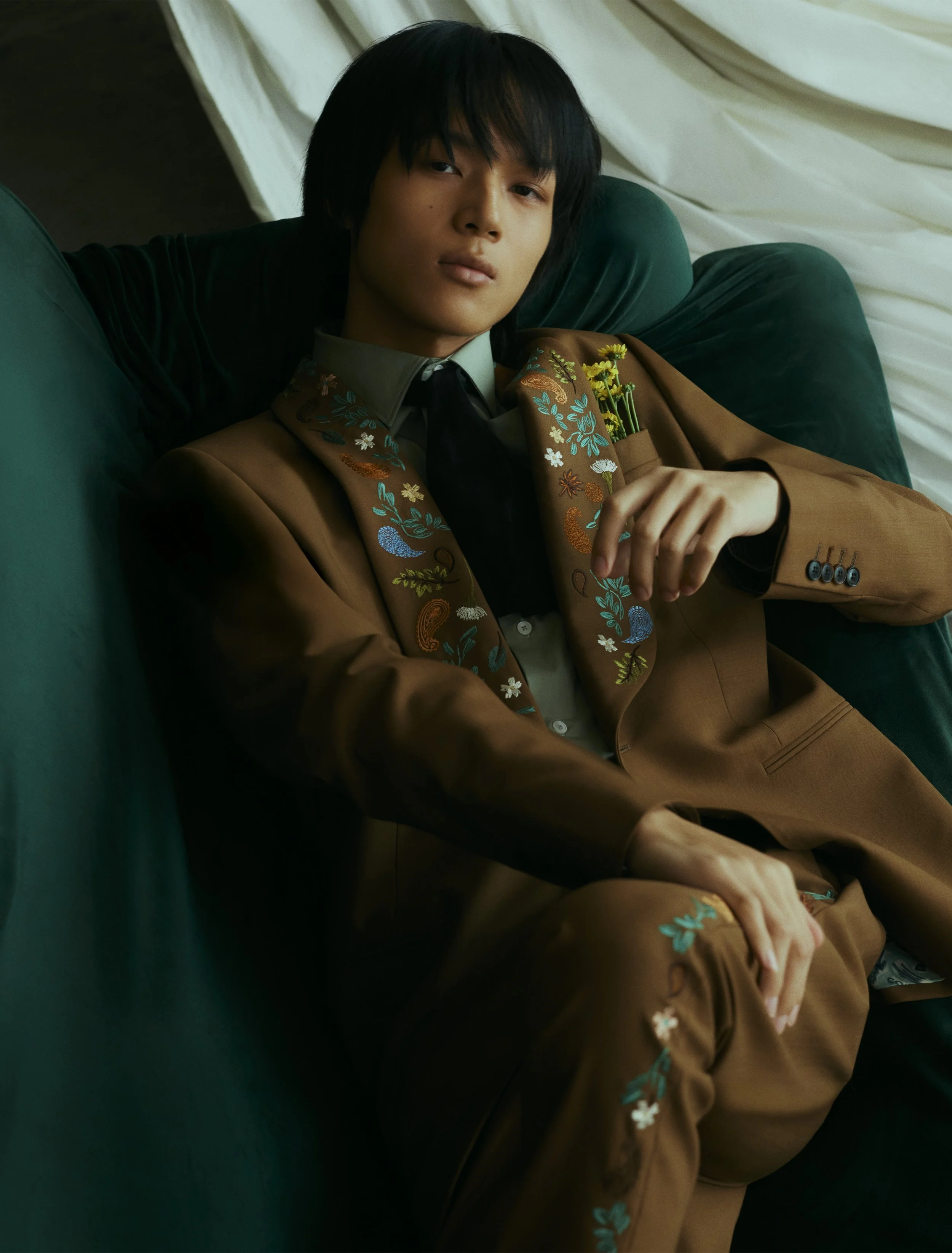A young person with black hair reclining on a green velvet sofa, wearing a brown suit with colorful embroidery and holding a small bouquet of yellow flowers.