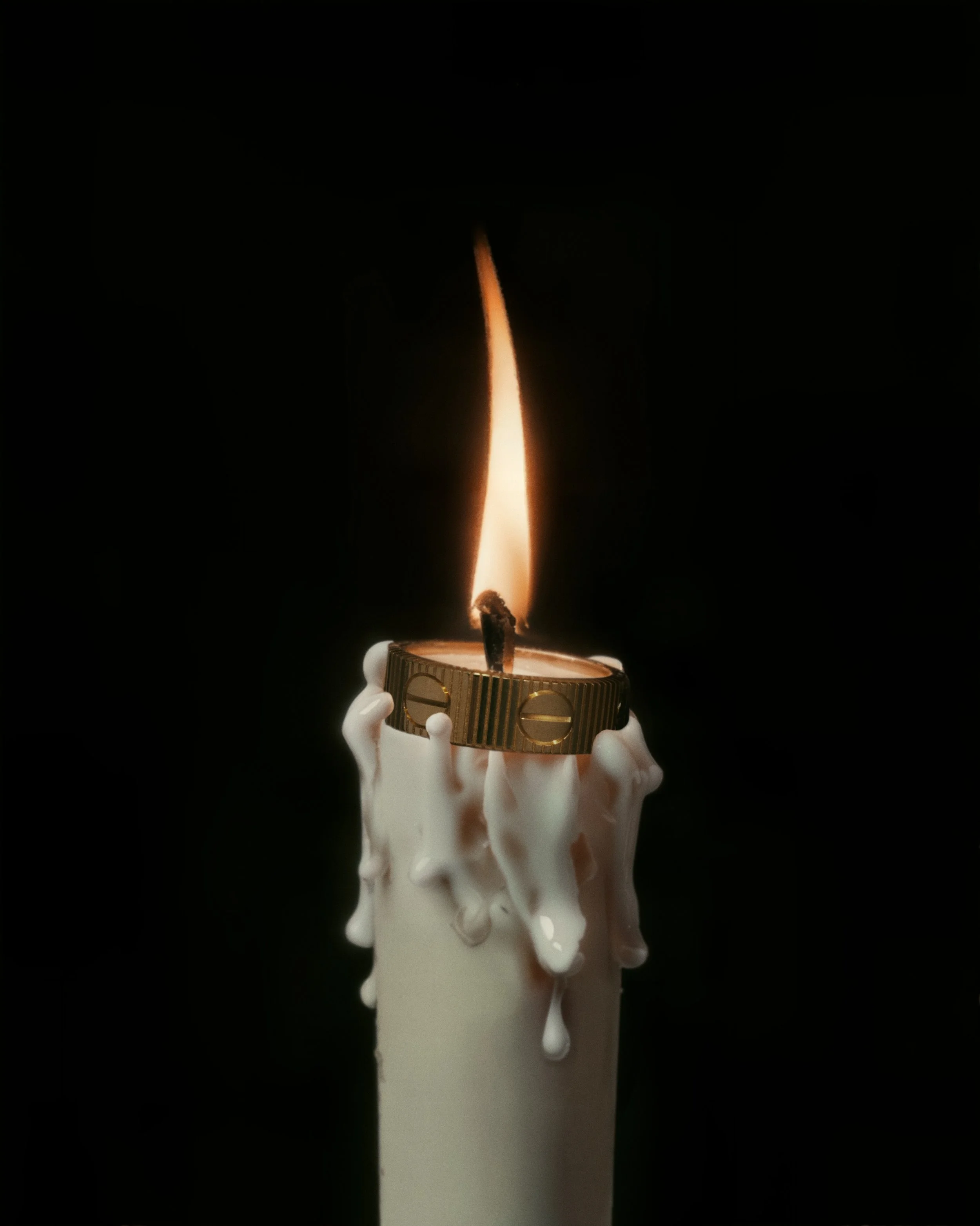 A lit candle with white wax melting and dripping down holds a gold ring-shaped object with engraved details in a dark background.