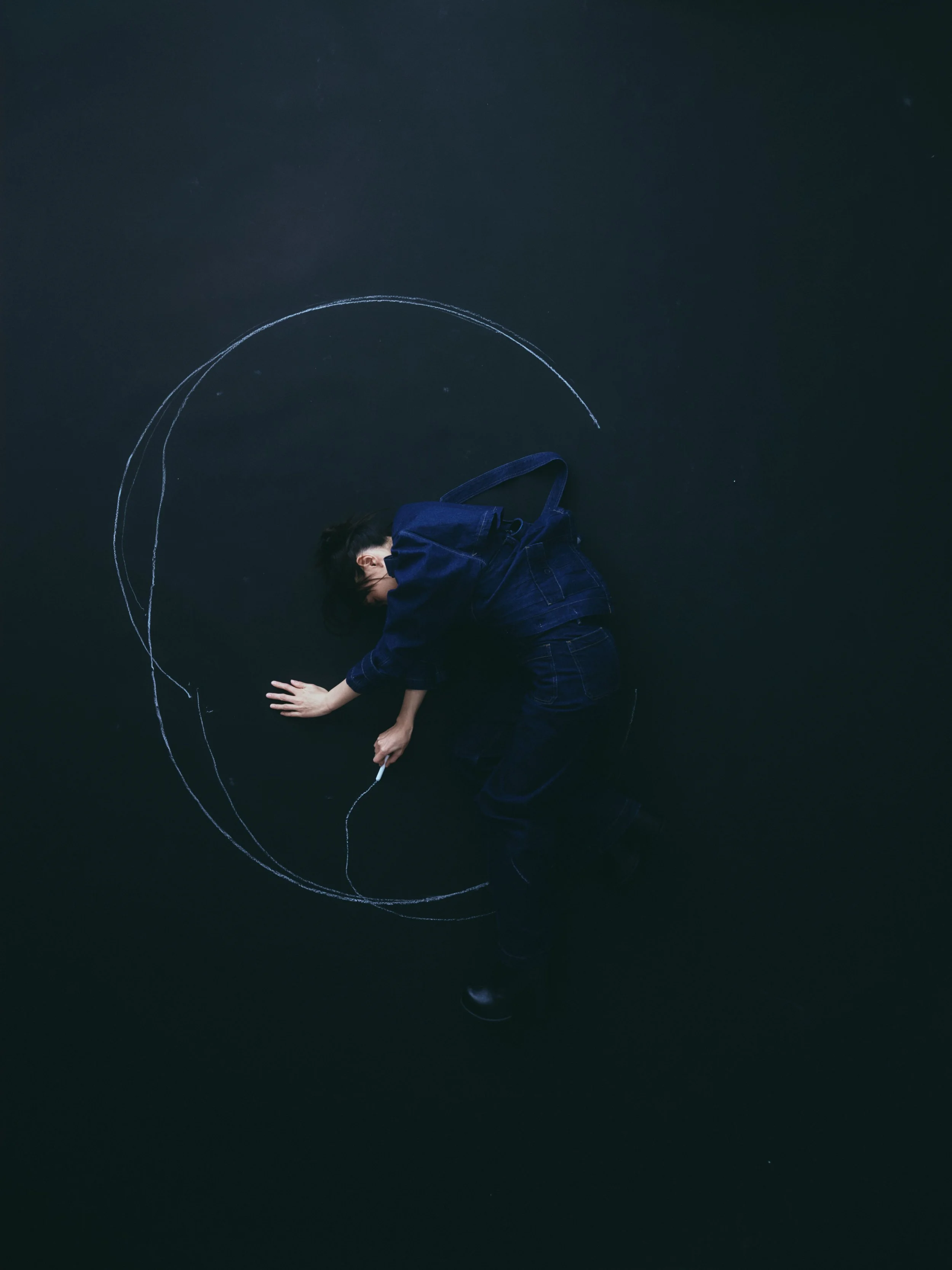 Ocean On, Person lying on the dark floor, drawing with white chalk to create a large circle.