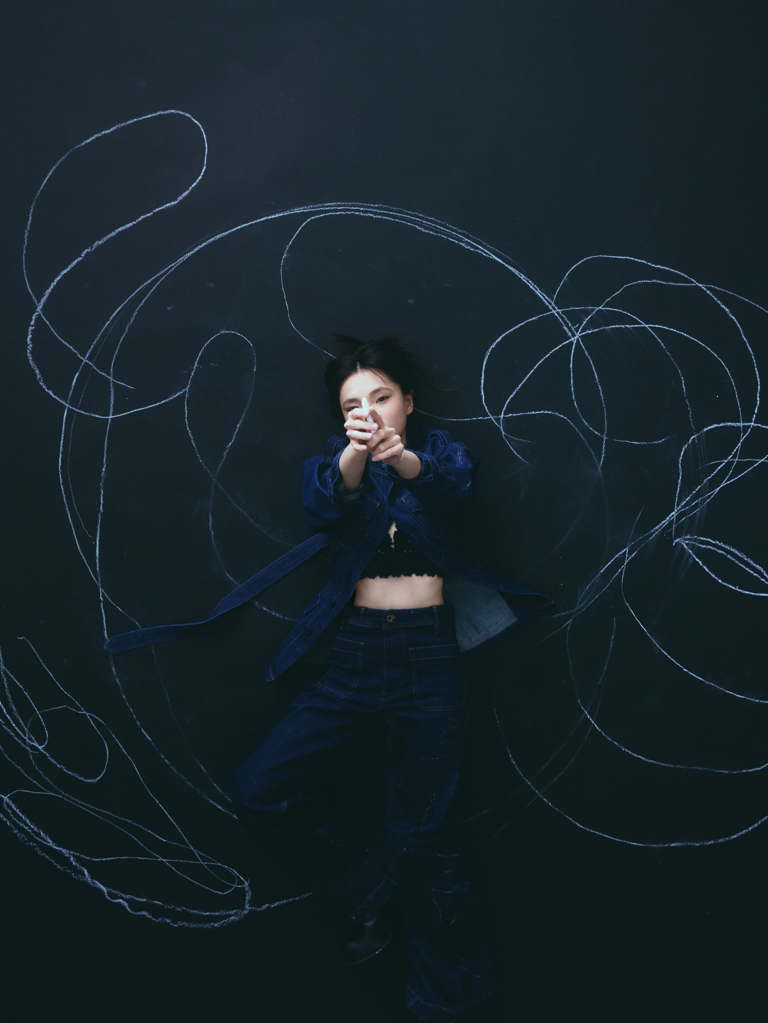 Ocean On,A young woman lying on a black floor with white chalk doodles around her, wearing a dark blue jacket and black pants, with her hands clasped in front of her face.