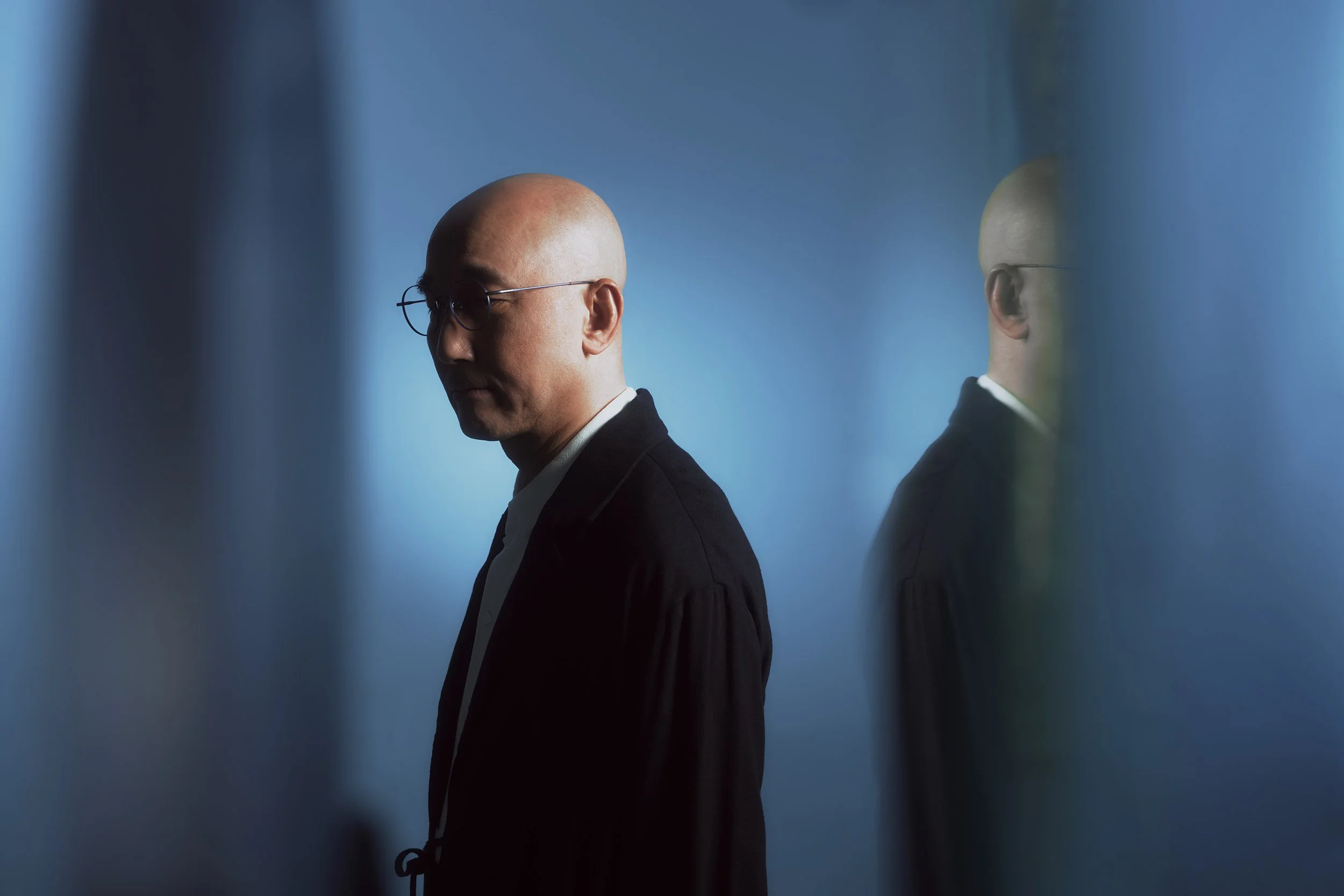 Lowell Lo, Bald man in glasses wearing a suit, standing with head bowed against a blue background, mirrored by his reflection.
