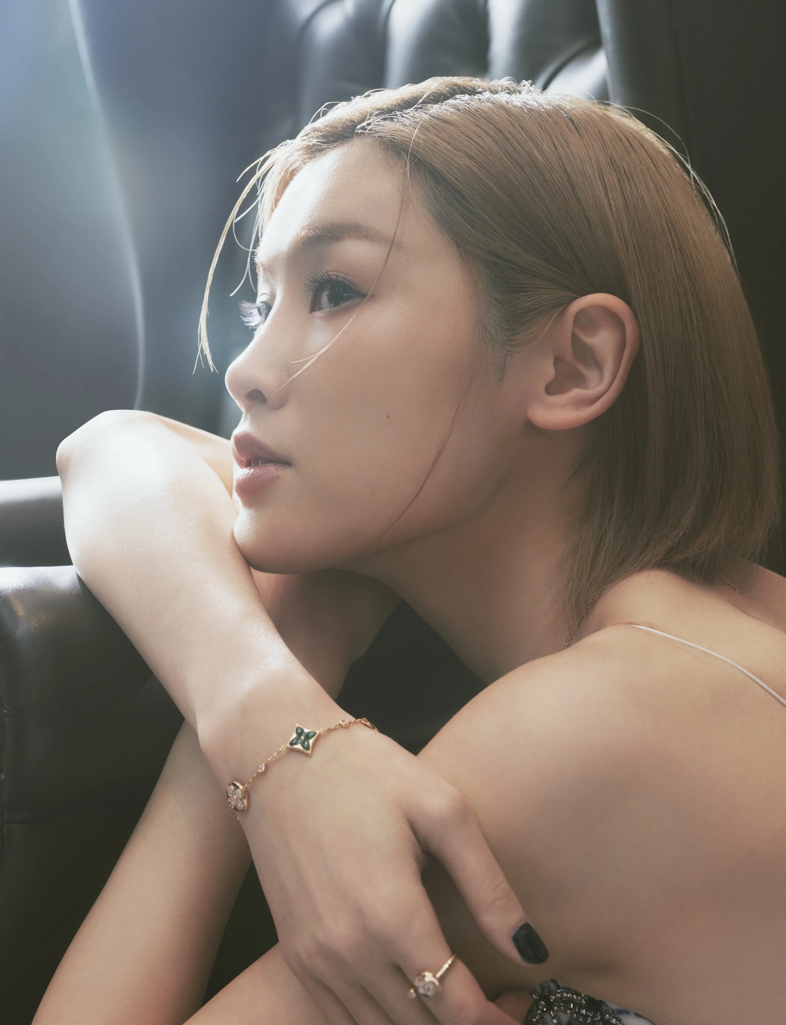 A close-up of a woman's face and upper body, with her head resting on her arm, revealing her eye, nose, and lips with makeup, wearing jewelry, and with light brown hair, sitting in a dimly lit environment.