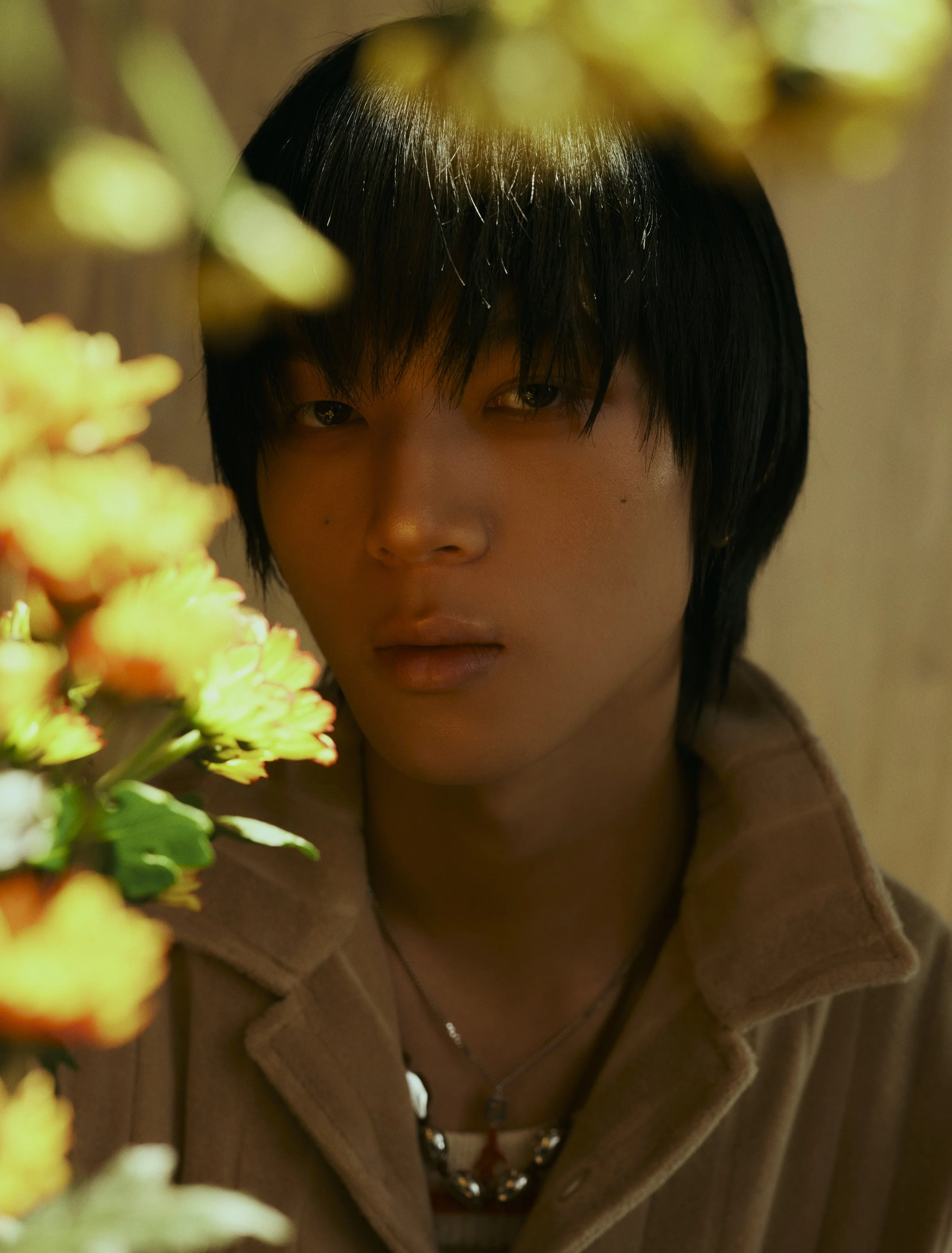 A person with short black hair and light skin wearing a brown fleece jacket and a silver necklace, partially obscured by yellow flowers in the foreground.
