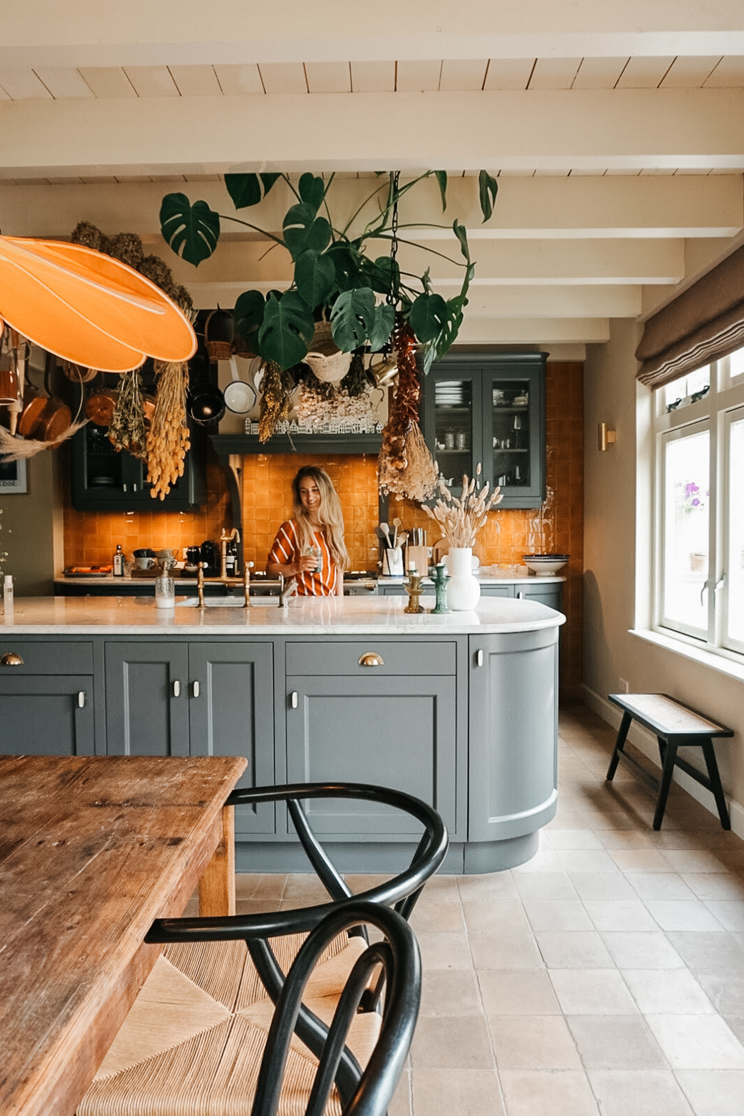 Een vrouw met lang blond haar in een keuken, aan een blauwe keukenkast. De keuken heeft een oranje betegelde muur en een tafel met zwartere stoelen. Boven de keuken hangt een groot groene plant en er hangen gedroogde bloemen.