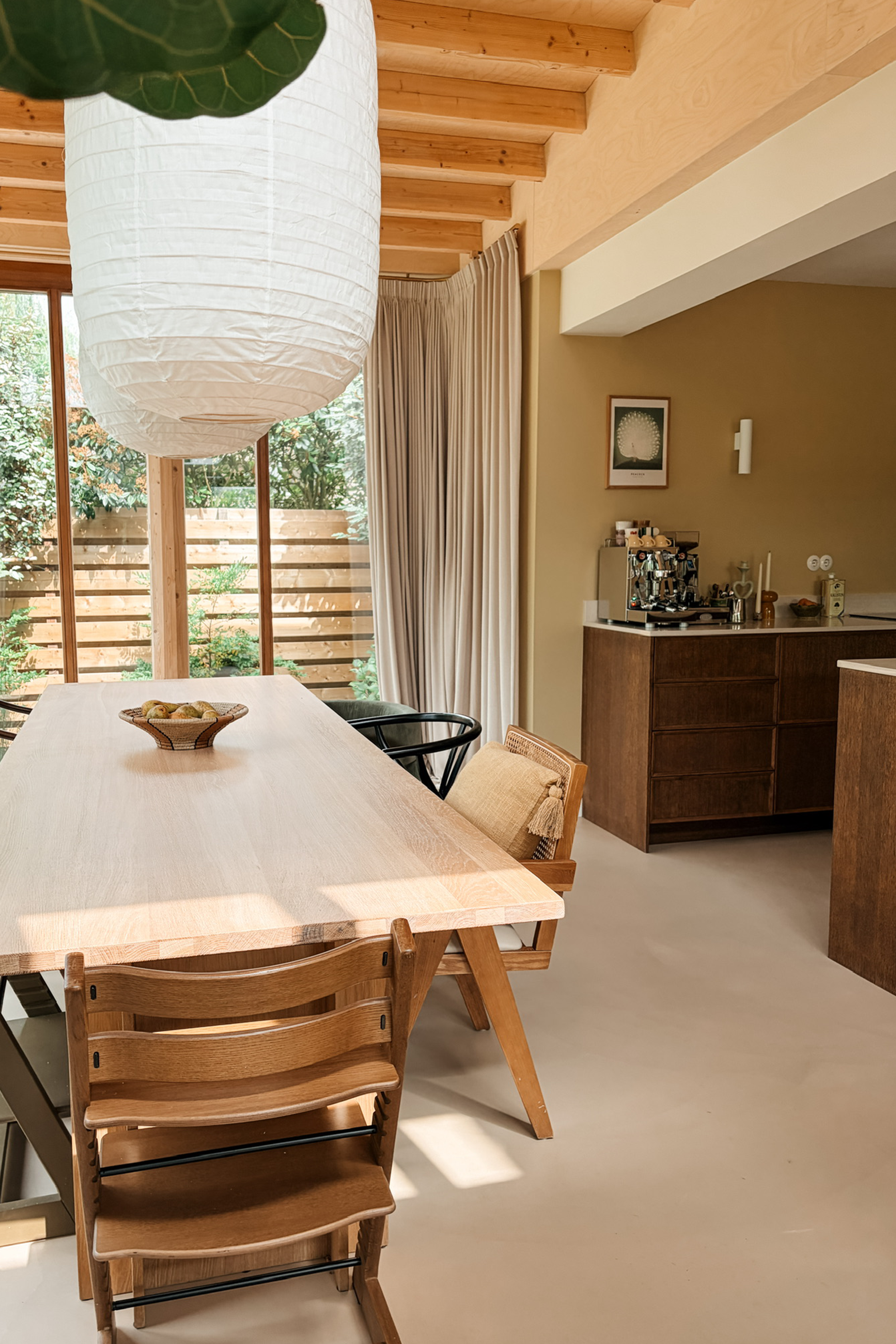 Binnenzichten van een gezellige kamer met een grote houten tafel, houten stoelen, een witte papieren lampenkap, grote ramen met beige gordijnen, een buffetkast met apparatuur en decoratie, en een wereldkaart aan de muur.
