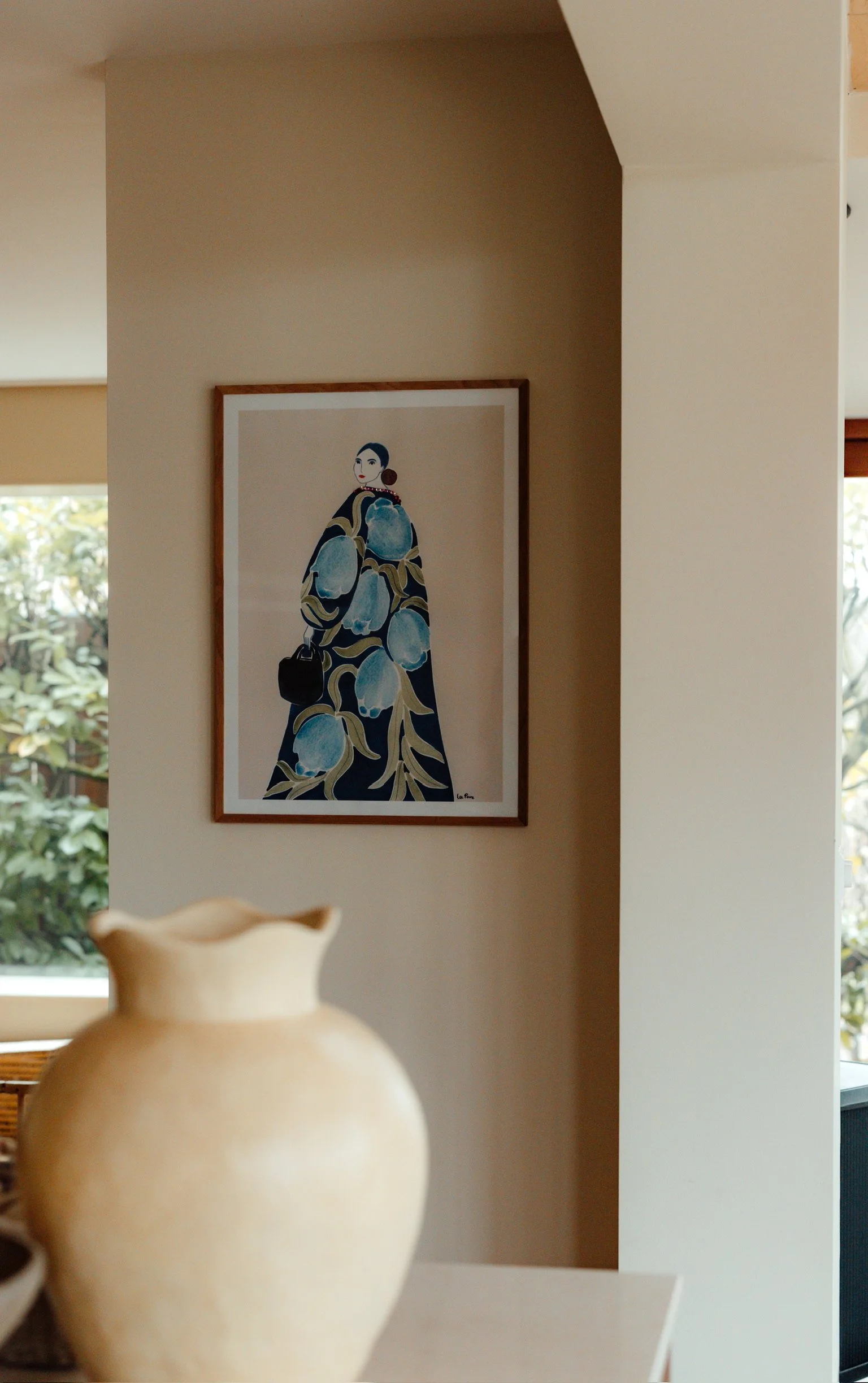 Een witte vaas op een tafel, en op de muur hangen schilderijen van een vrouw in een blauwe jurk met een zwart tas.