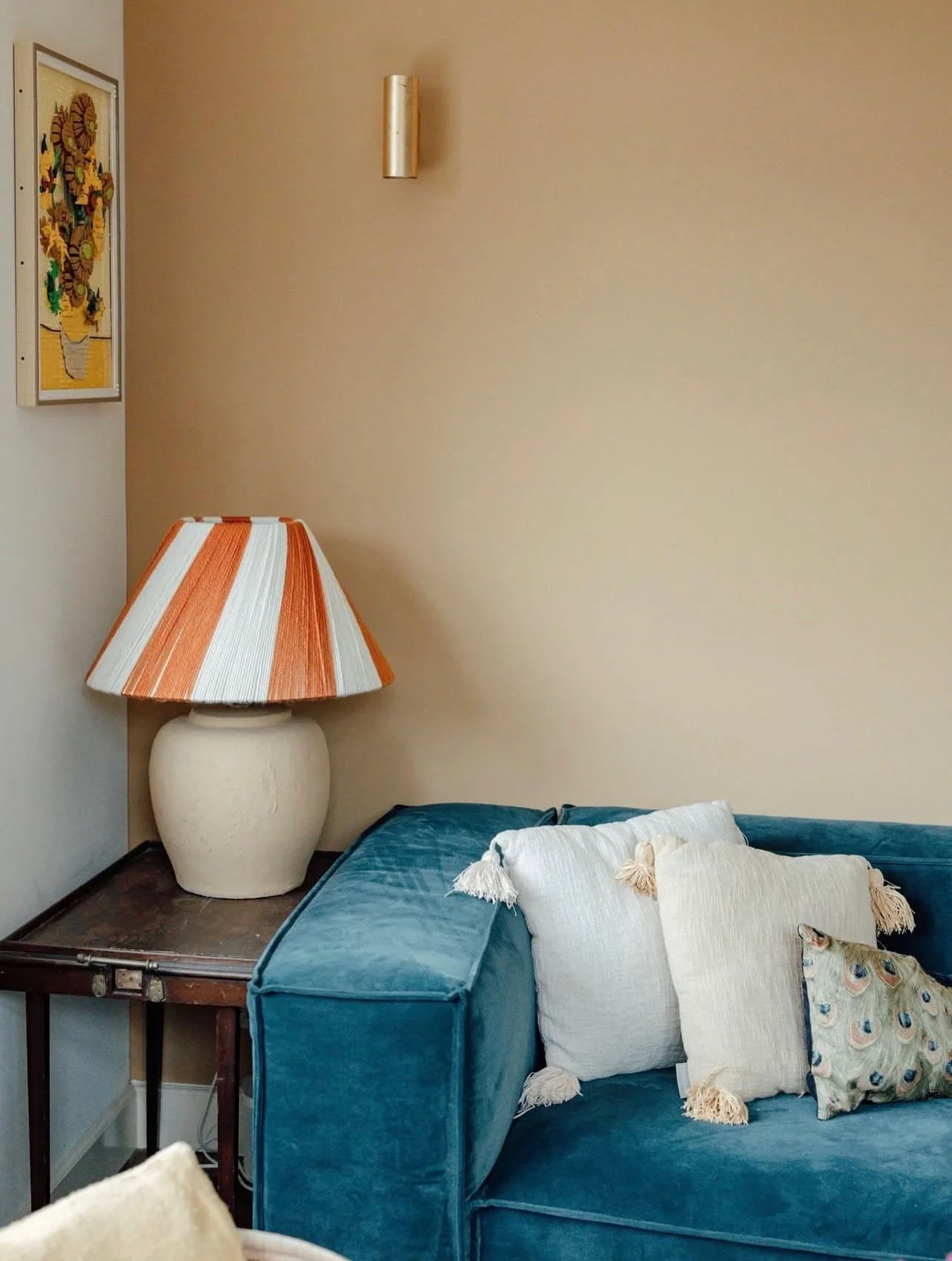 Een beige muur met een wandlamp, een schilderij en een salontafel met een grote keramieken lamp en een bank met kussens.