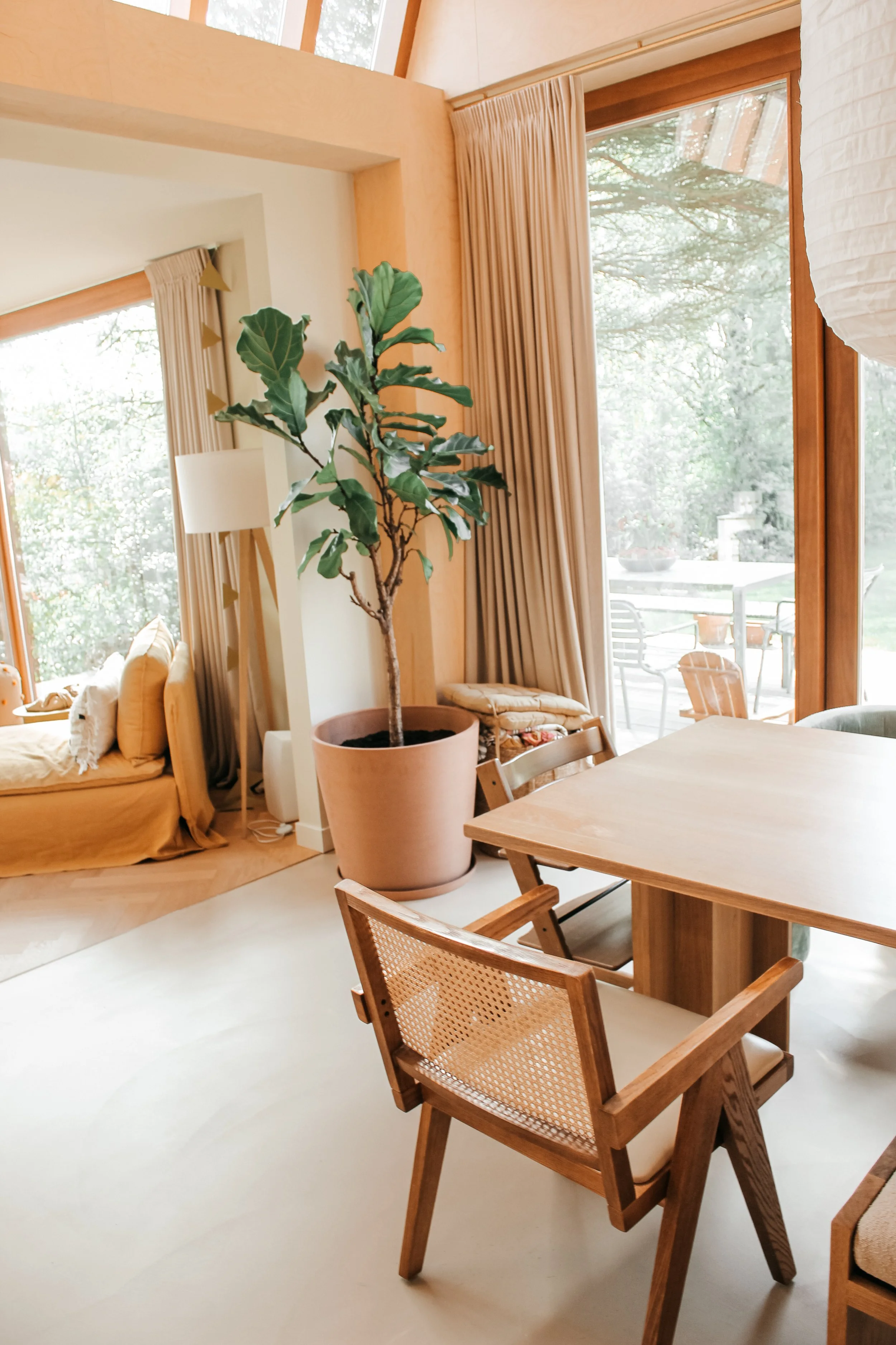 Een woonkamer met grote glazen deuren die uitzicht bieden op een tuin. Er staat een grote groene plant in een roze pot, een houten tafel en houten stoelen. De ruimte is licht en gezellig.