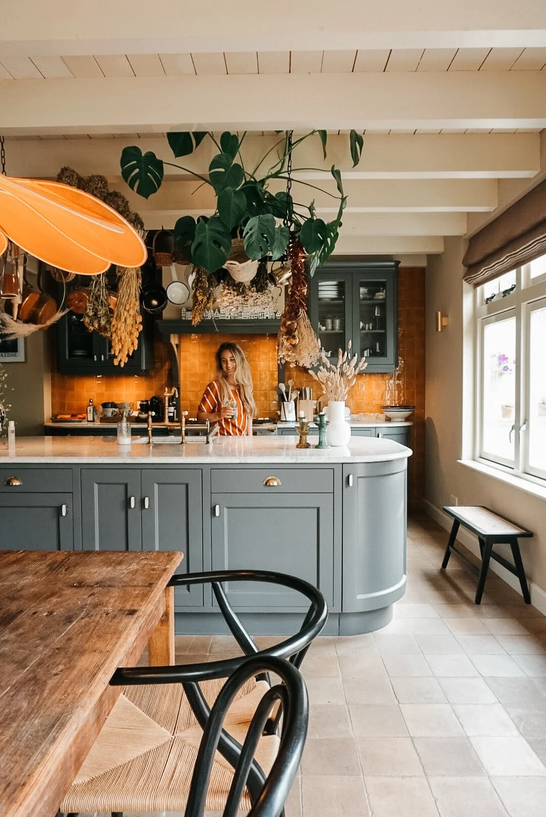 Een moderne keuken met een blauwe keukenkast en een wit aanrecht. Een vrouw met lang haar in een rood-wit gestreepte top staat achter het aanrecht, drinkend uit een glas. Er hangen gedroogde bloemen en een grote plant aan het plafond. De keuken heeft een grote raam met uitzicht op buiten, een kleine bankje onder het raam, en een houten eettafel met een zwart rugleuningstoel.