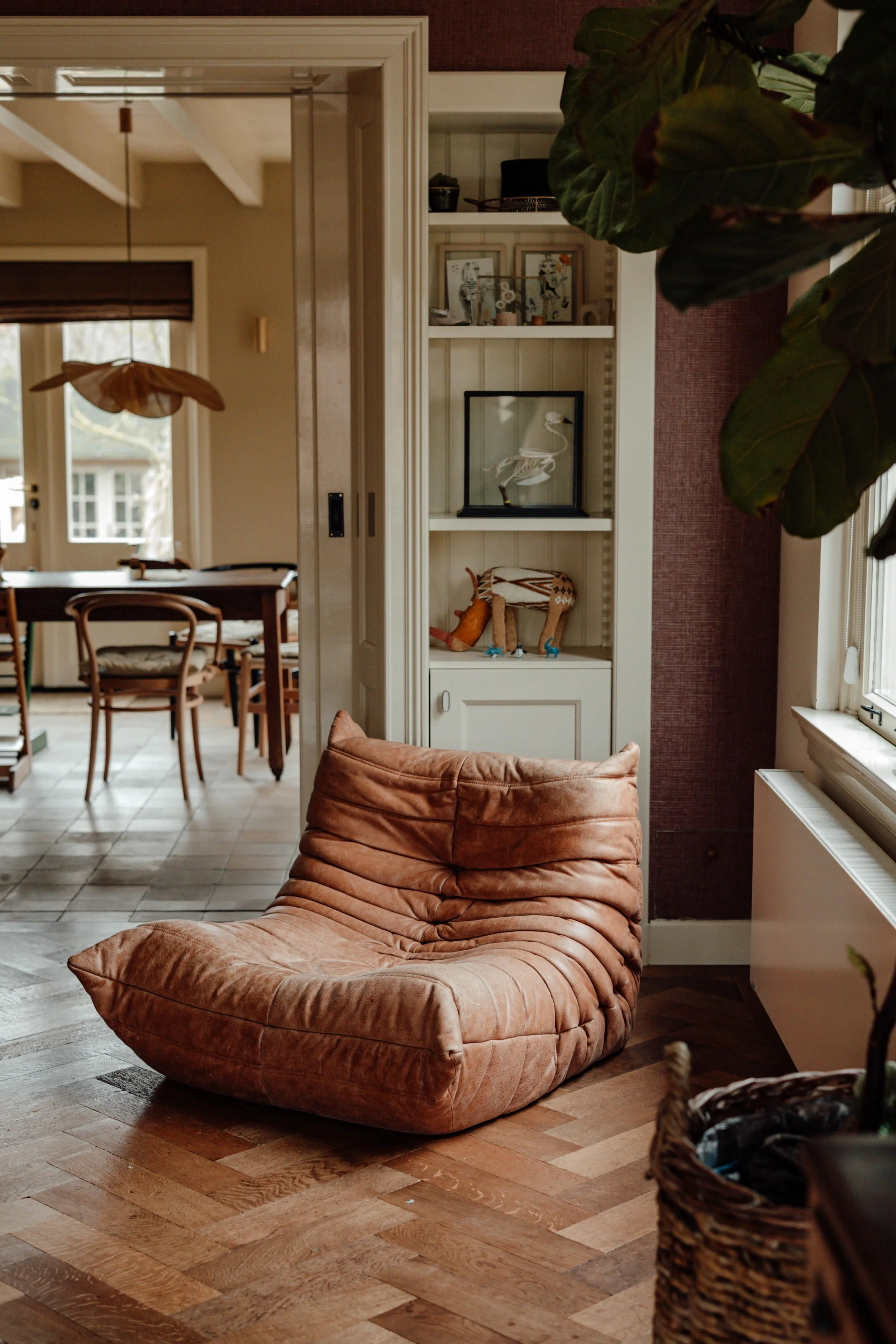 Een gezellige woonkamer met een grote, roze, zachte fauteuil in de voorgrond, een witte boekenkast met decoratieve items en een vogelbeeld, een raam met witte kozijnen en een gordijn, een houten eettafel met stoelen in een aangename lichtinval.