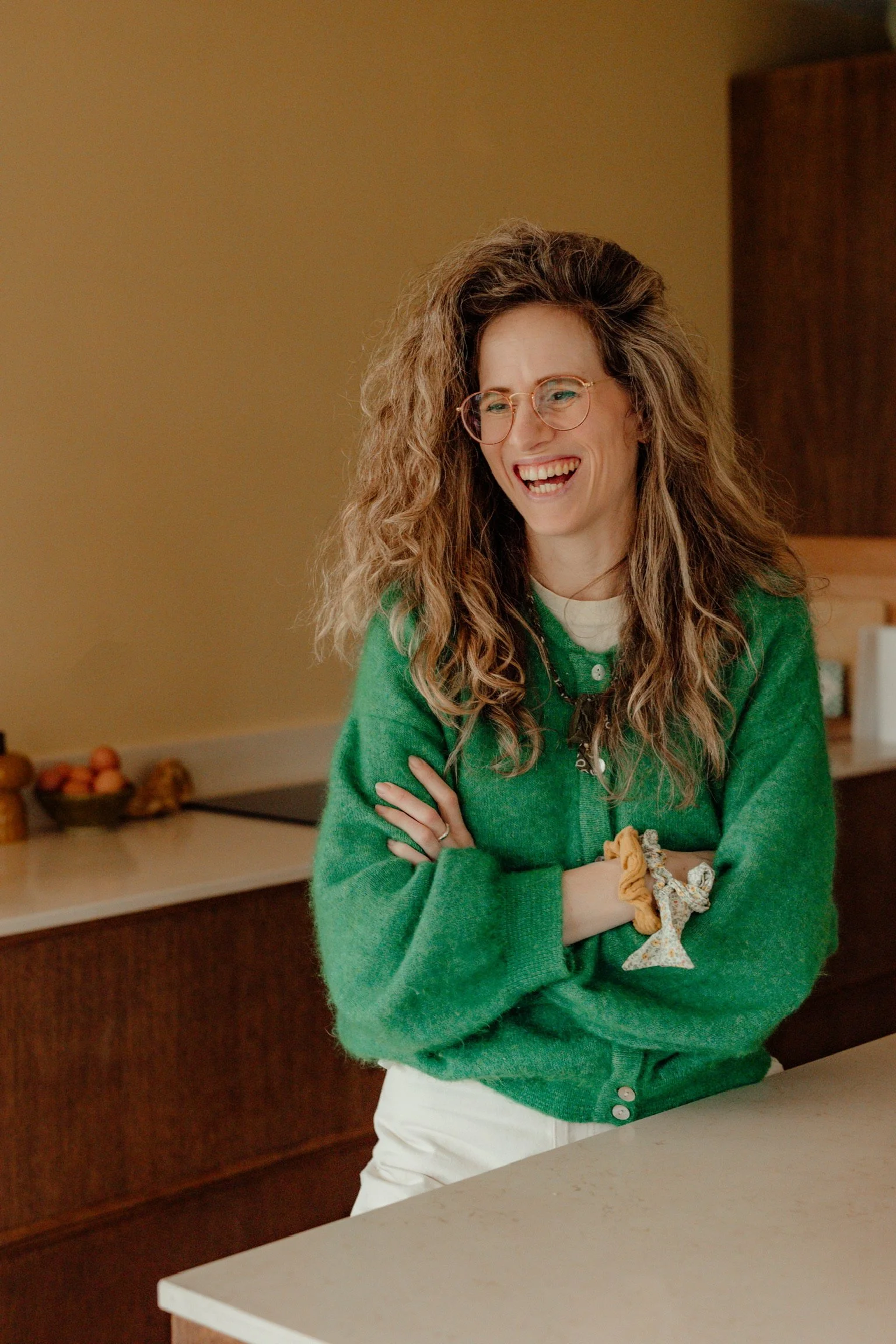 Vrouw met lang, gekruld haar en brillen, lachend terwijl ze in een keuken staat. Ze draagt een groene trui en witte broek.