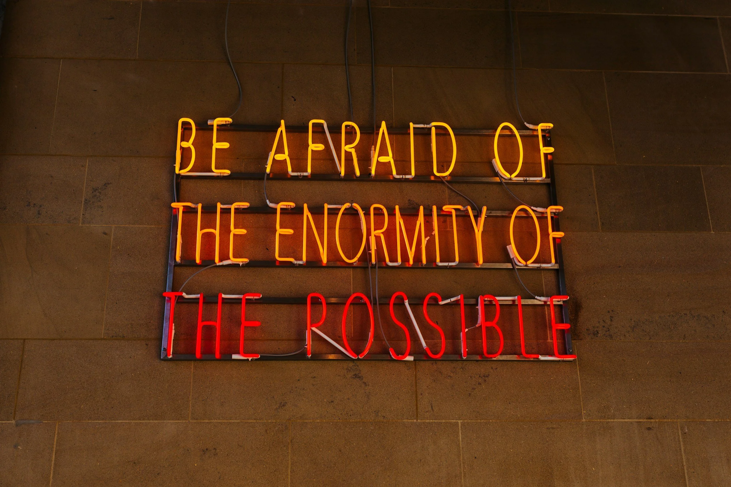 Neon sign on a wall reads 'BE AFRAID OF THE ENEMY OF THE POSITIVE' with the top row in yellow and the bottom row in red.