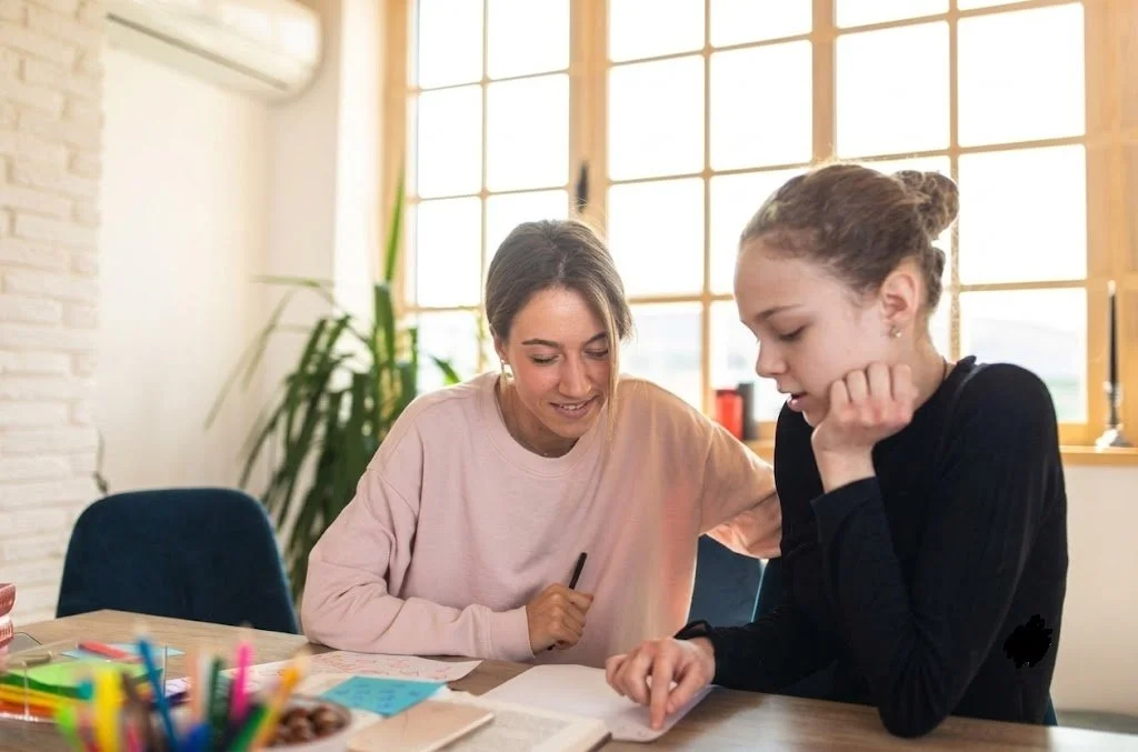 Young lady receiving speech and language support at a table, surrounded by colorful pens and papers in a bright, cozy room with large windows.