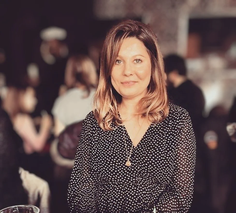 A woman with shoulder-length light brown hair, wearing a black dress with white polka dots and a heart pendant necklace, smiling in a dimly lit social setting.