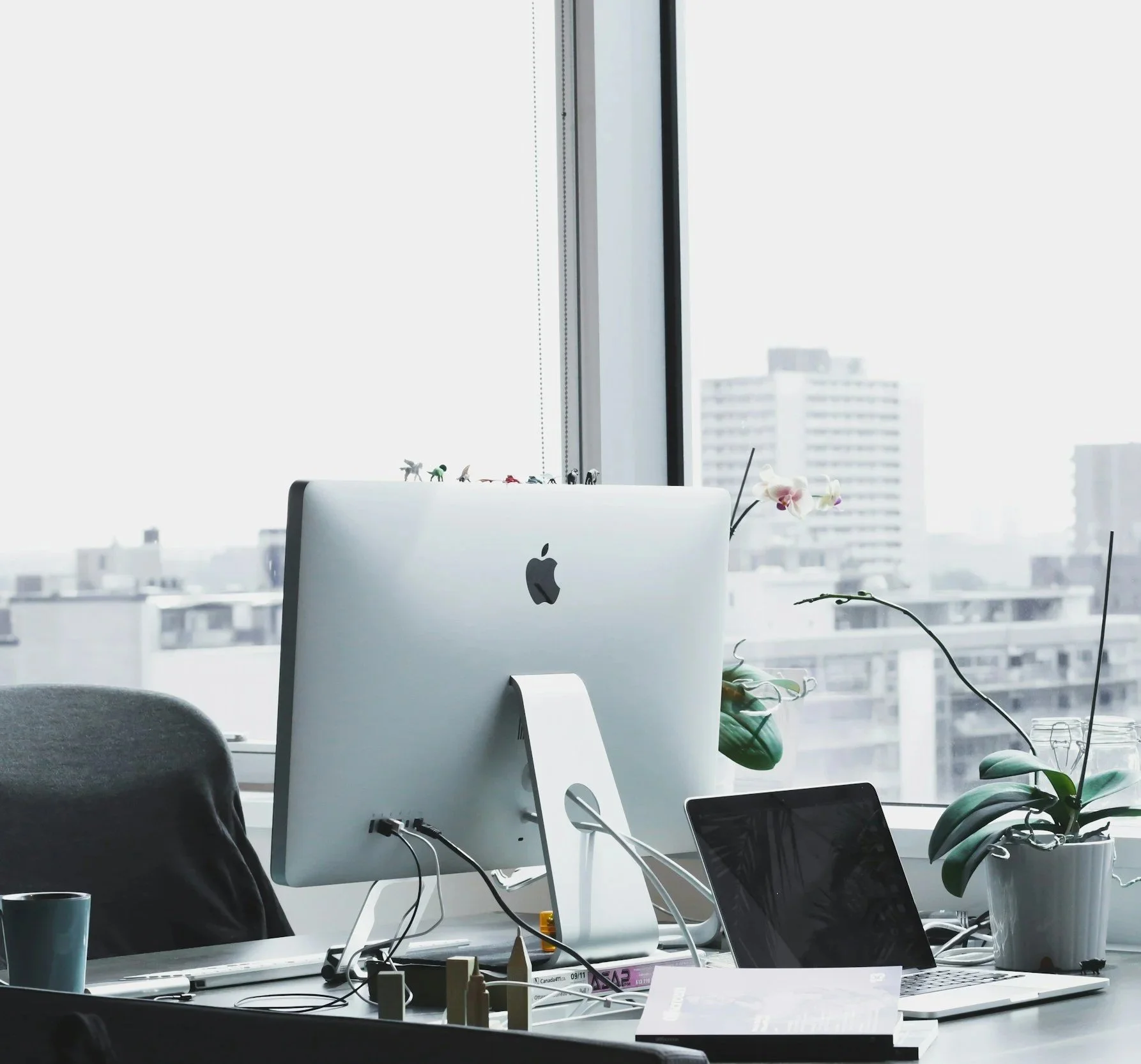 A modern office workspace with a computer monitor, laptops, and various office supplies on a desk, set against large windows overlooking city buildings.