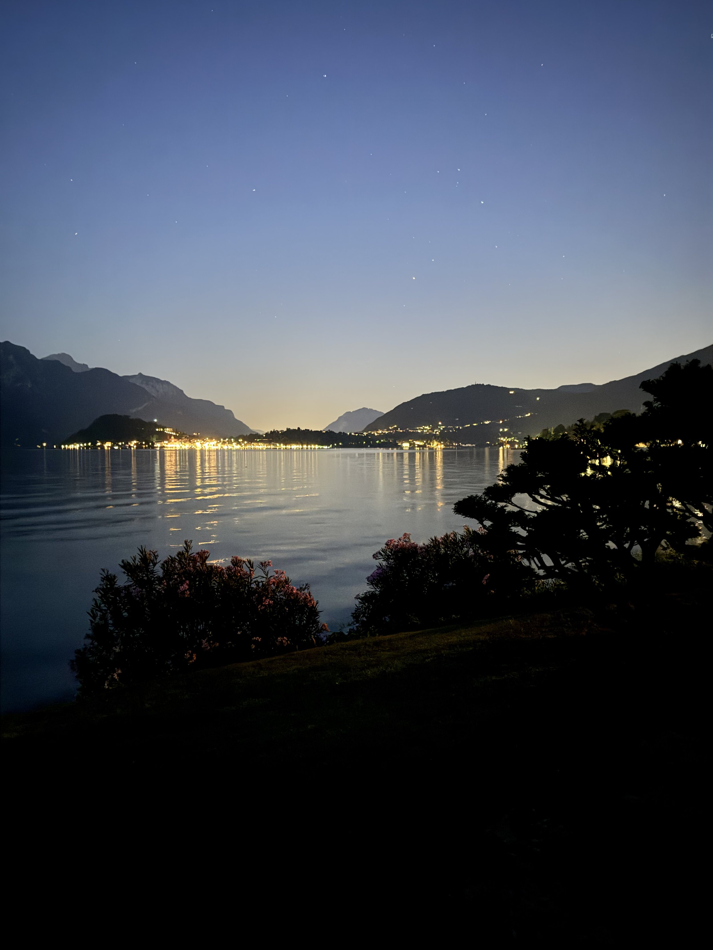 Nighttime landscape with a body of water reflecting lights from a town or village on the shore, surrounded by mountains under a clear sky with visible stars.