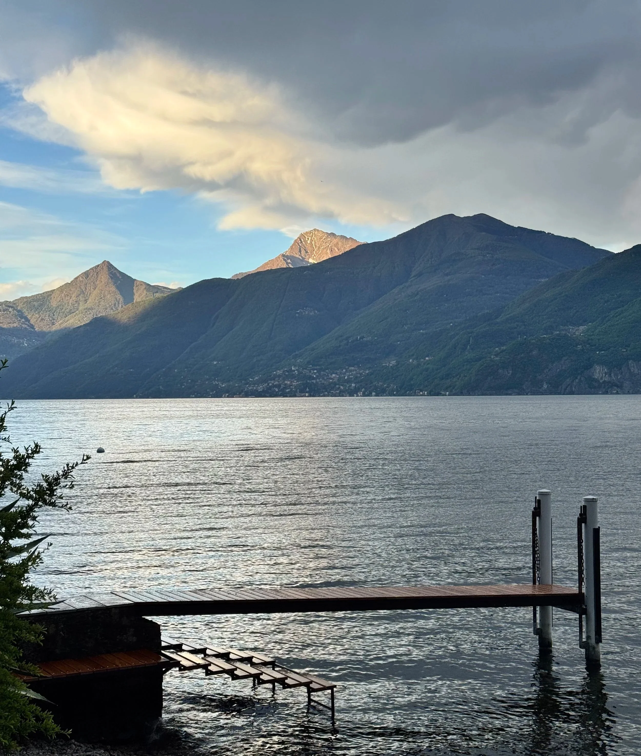 A lakeside scene with a wooden dock extending into the water, mountain ranges in the background, cloudy sky, and lush greenery on the shore.