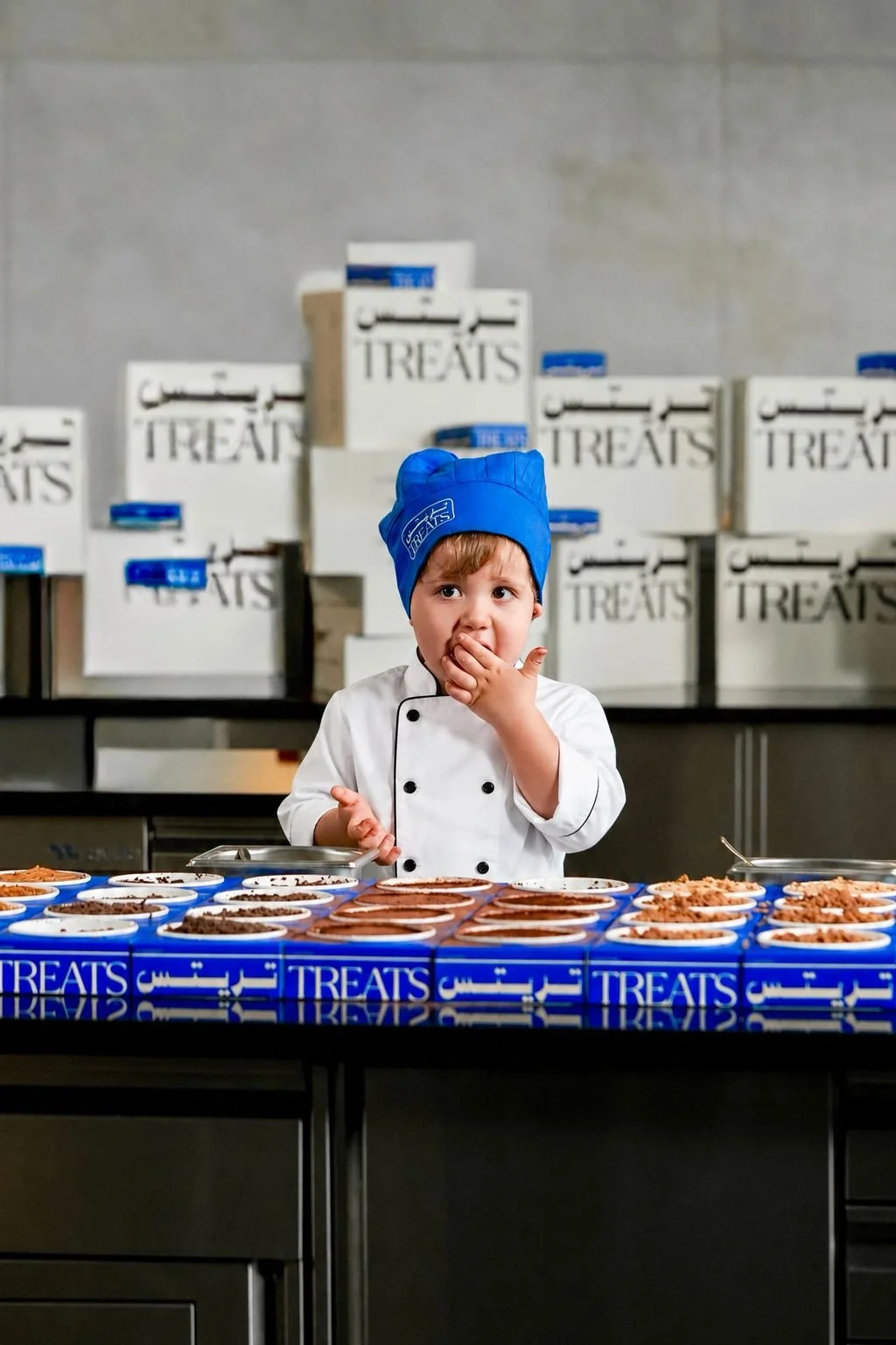 A young boy dressed as a chef in a white coat and a blue chef hat, tasting treats at a display in a kitchen with boxes labeled 'treats' in the background.