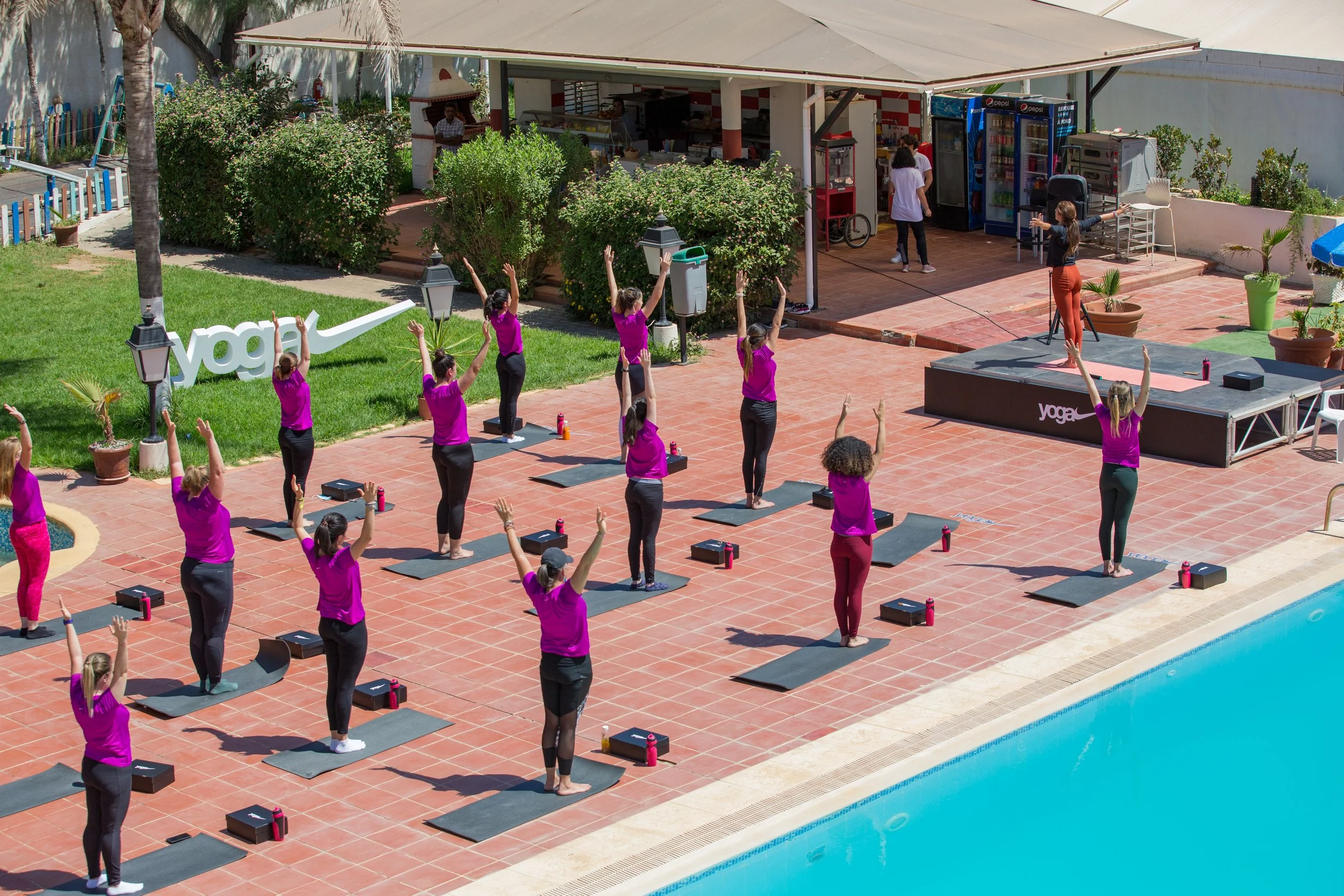 A group of women in purple shirts and black pants participating in a yoga class outdoors near a swimming pool. They are standing on yoga mats with their arms raised, following the instructor who is standing on a stage.
