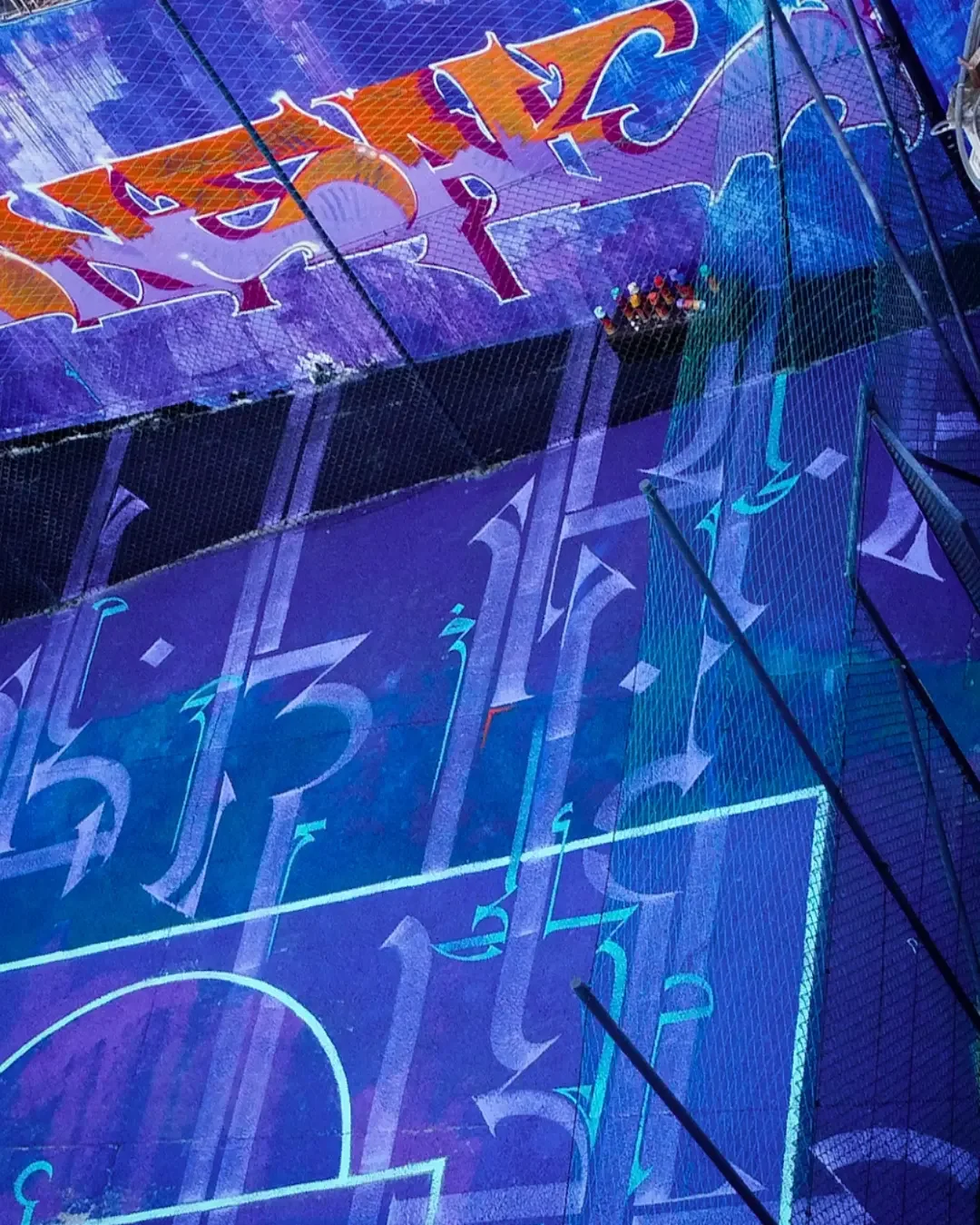 Colorful neon lighting and markings on a sports court, including part of a soccer field and a basketball court, viewed through a sporty fence.