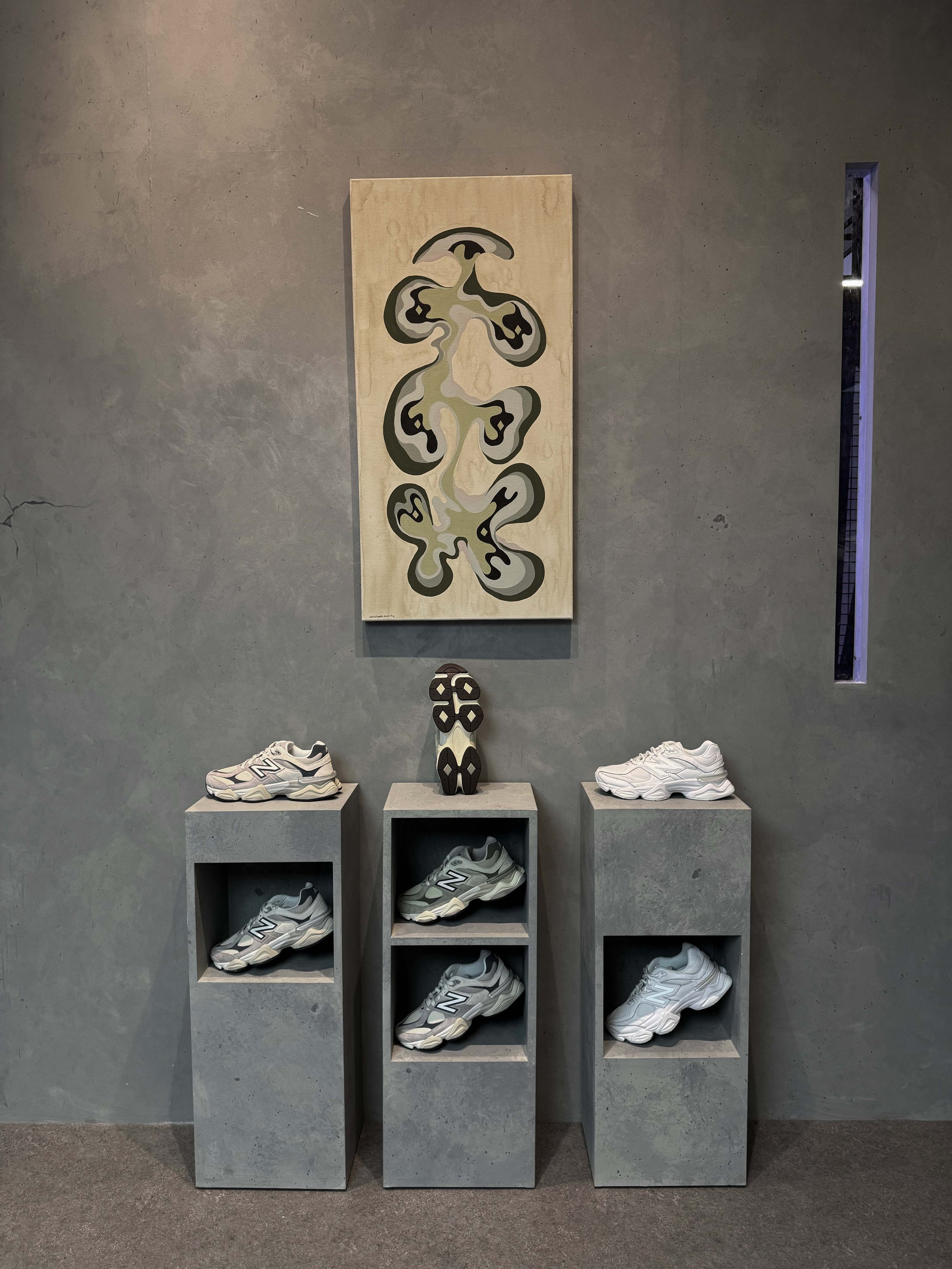 Display of New Balance sneakers on grey concrete pedestals and shelves against a grey wall with abstract artwork and narrow window.