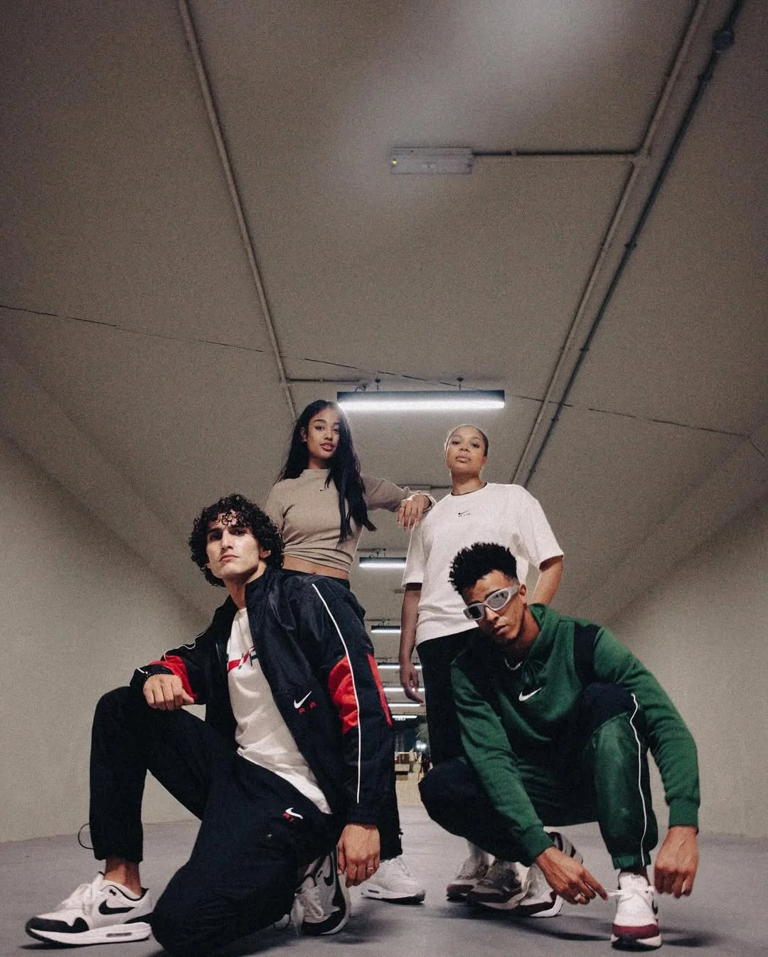 Four young adults posing in an underpass, dressed in sporty casual clothing and sneakers.