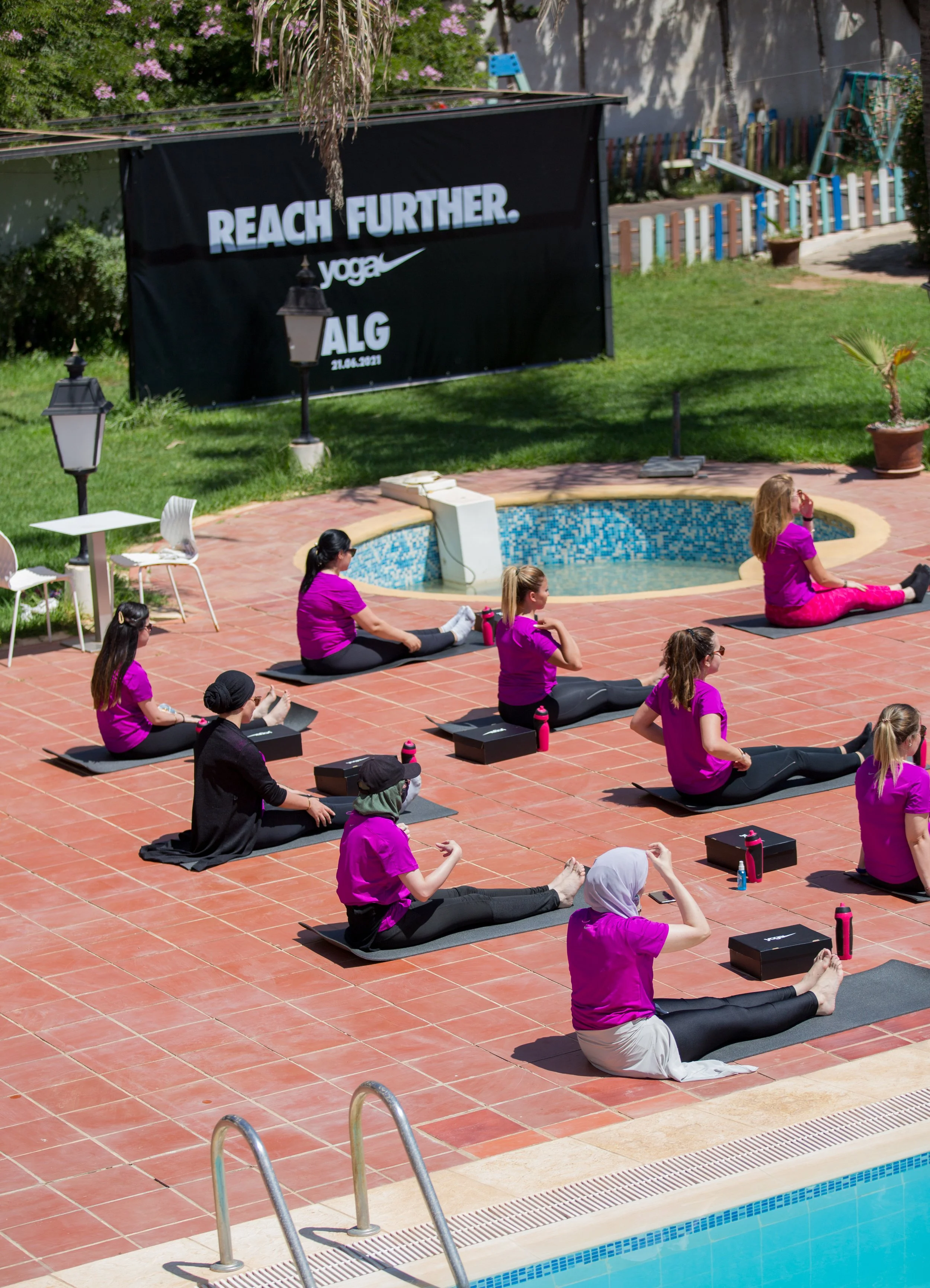 A group of people practicing yoga outdoors on a poolside patio, sitting on black yoga mats in a cross-legged position, with some wearing purple shirts and others in black or grey. There are water bottles and small boxes next to some participants, and a benefit or promotion banner in the background with the words 'REACH FURTHER. yoga ALG 21.06.2021'.