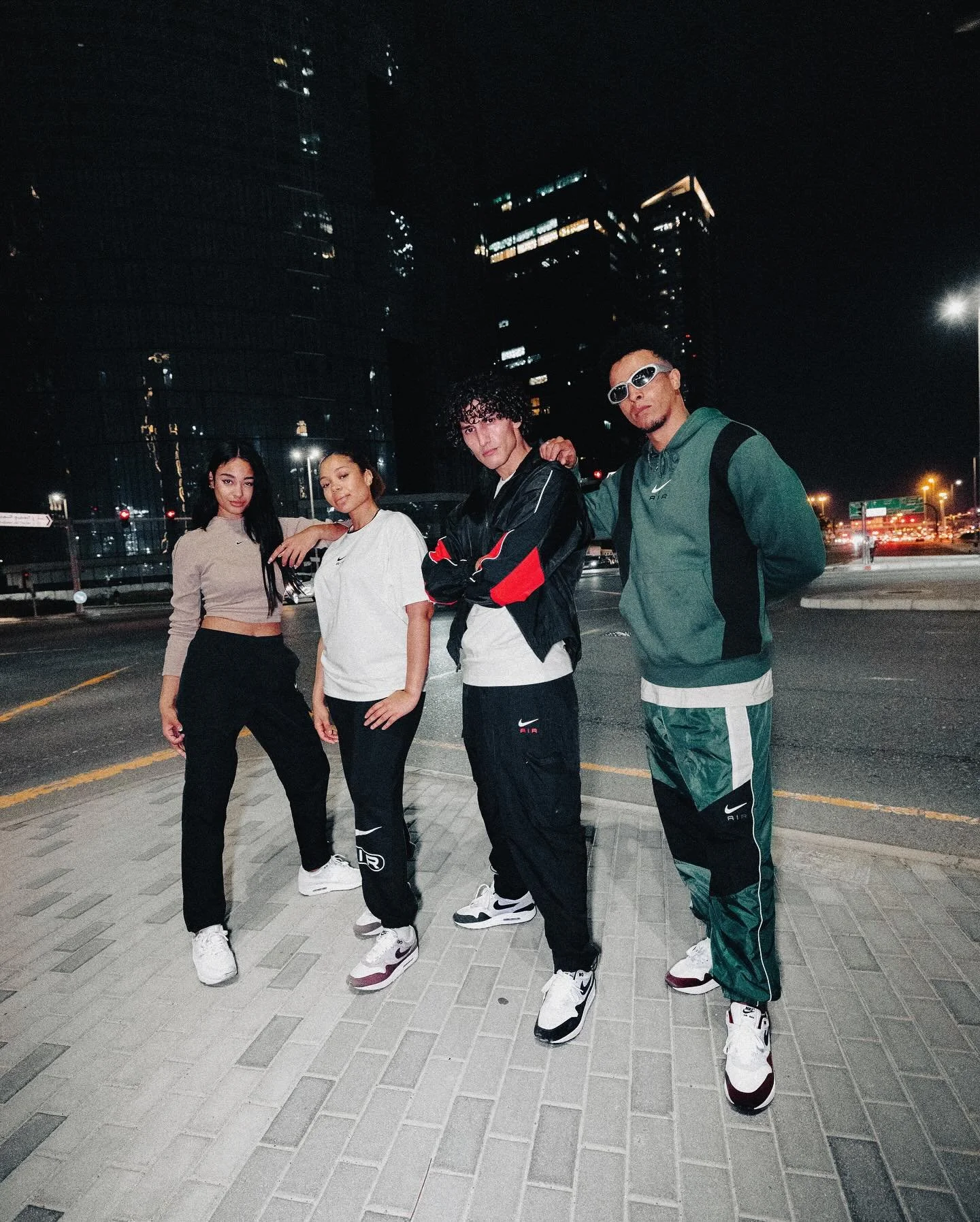 Group of four young adults posing on a city street at night with tall buildings in the background, dressed in athletic streetwear and sneakers.