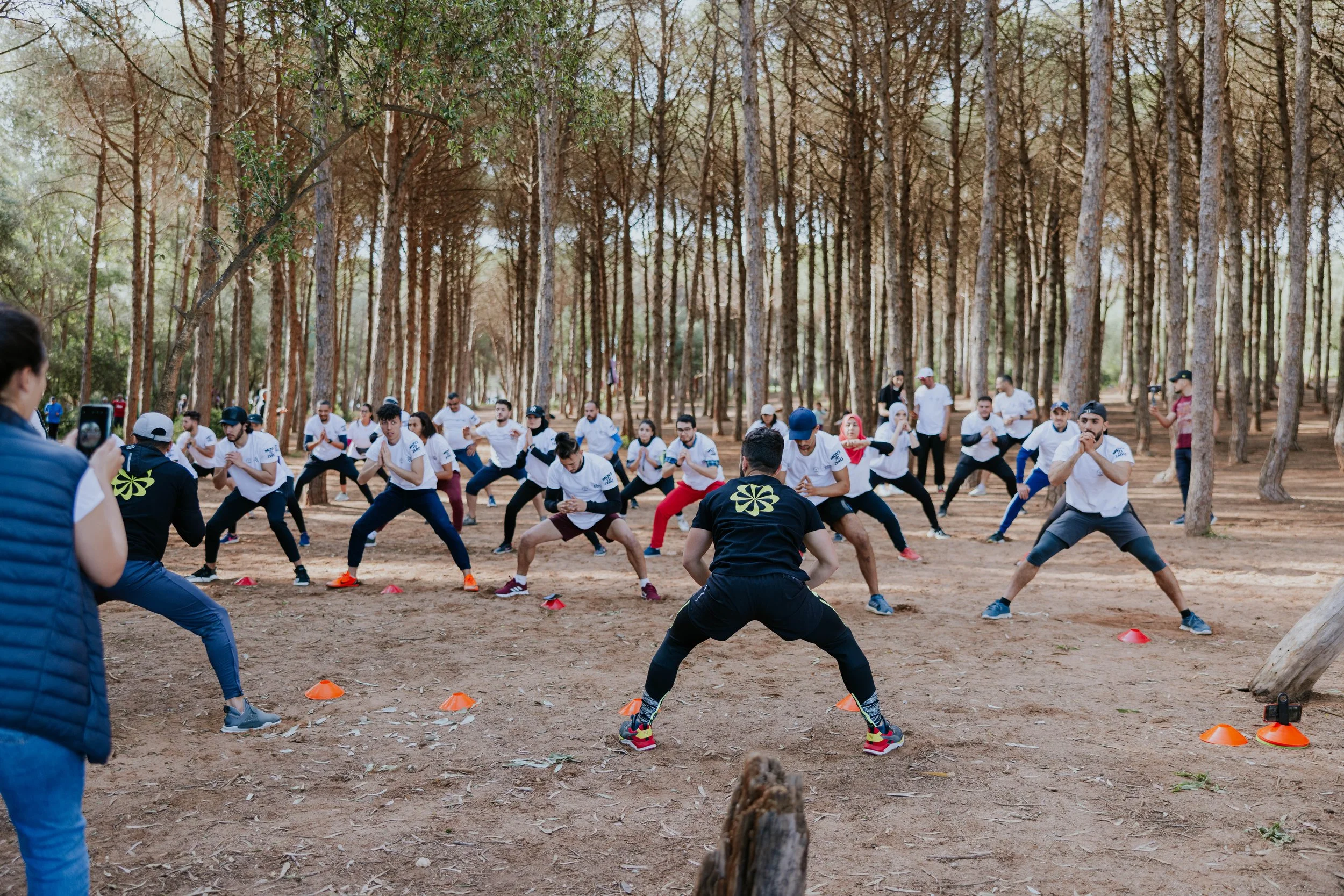 A group of people participating in an outdoor fitness class or training session in a forested area, with cones set up on the ground for exercises.