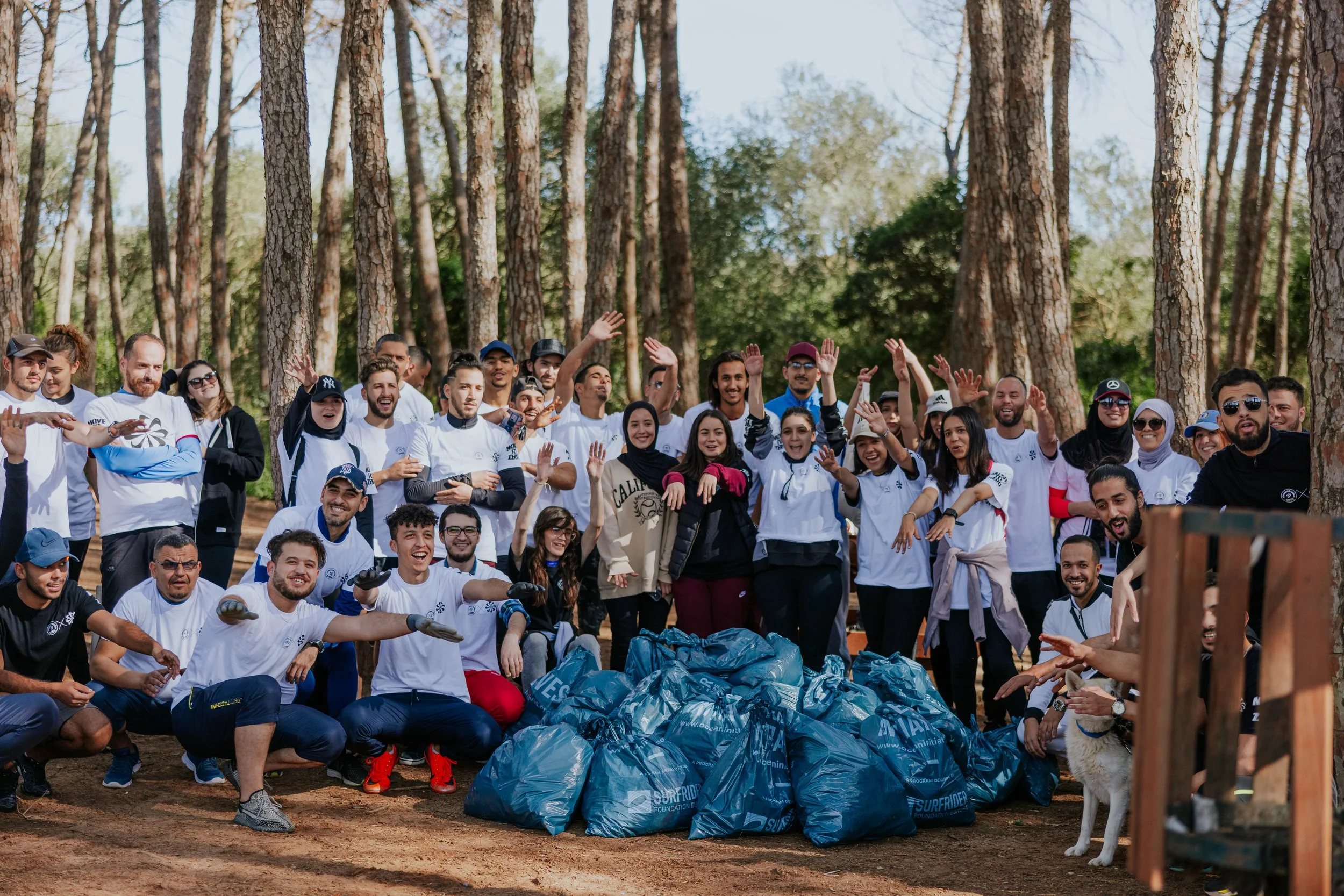 A large group of people gathered outdoors in a forest, participating in a volunteer activity. They are standing and sitting around, smiling and waving, with many wearing white T-shirts. There are blue trash bags filled with collected litter in the foreground, and a dog is present on the right side of the image.