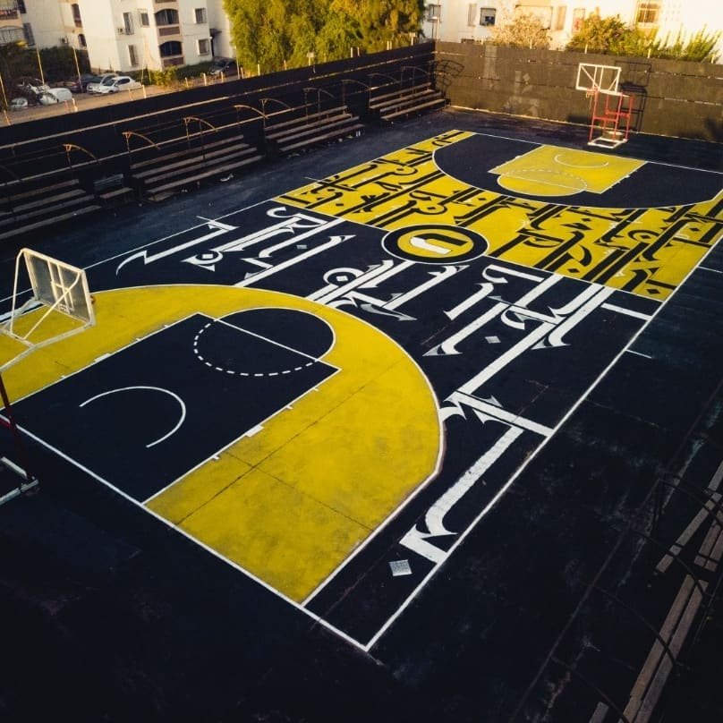 A basketball court with black, yellow, and white markings, including two hoops, surrounded by seating and trees.
