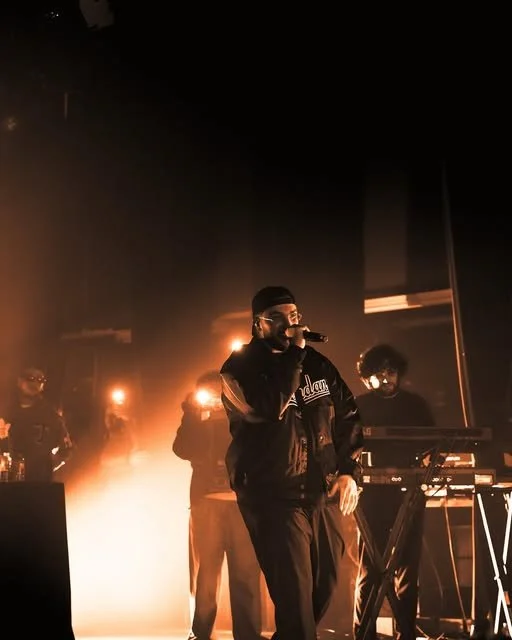 A rapper performing on stage with a microphone, wearing a black jacket and hat, surrounded by backup dancers or musicians, stage lights creating a warm glow.
