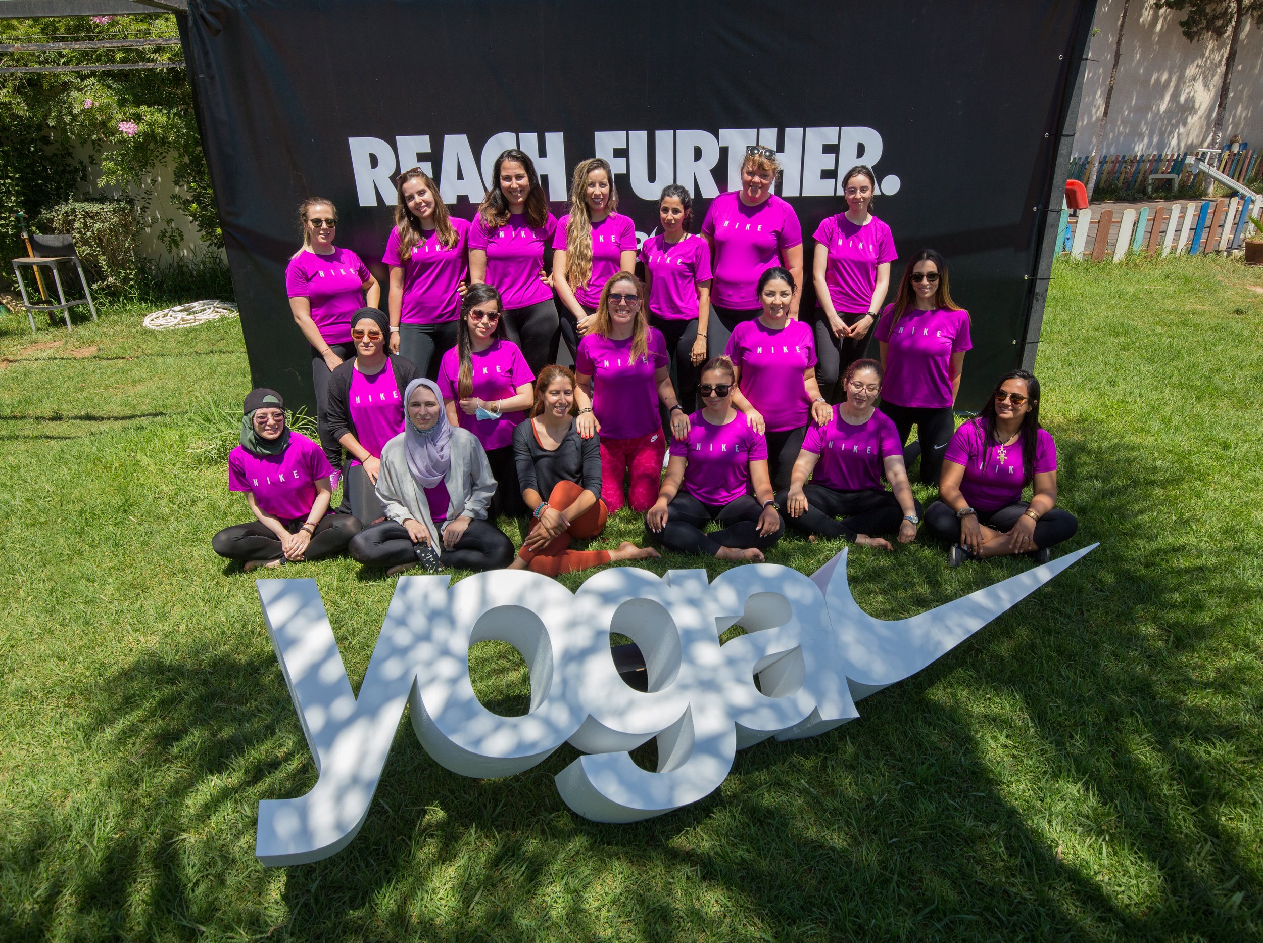 Group of women wearing pink Nike shirts, posing outdoors in front of a large black banner that reads 'PEACH FUETHER.' They are smiling and standing on grass, with some seated in front. There is a decorative white sign with partially visible letters on the ground.