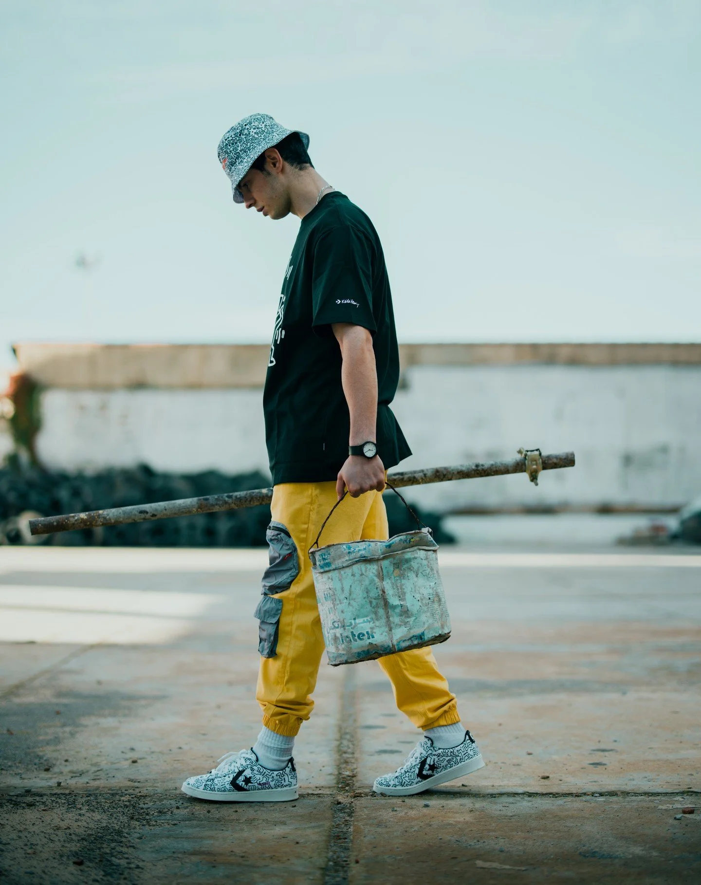 A young man wearing a patterned bucket hat, black t-shirt, yellow pants, and Nike sneakers with black and white patterns, holding a weathered metal bucket, walking outdoors near a construction site with a metal pipe and a white wall in the background.