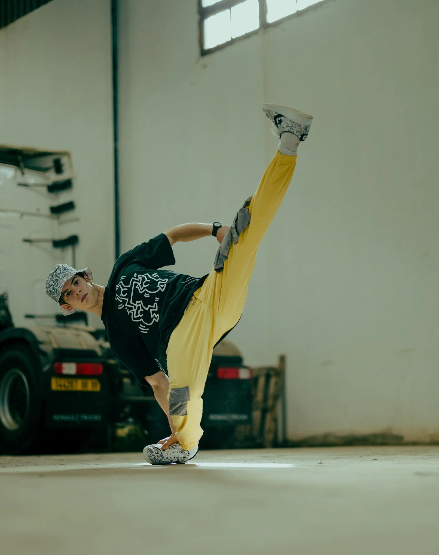 A young man in a black t-shirt, yellow pants, and a patterned cap performing a one-handed handstand in an indoor space with a plain wall and a car in the background.