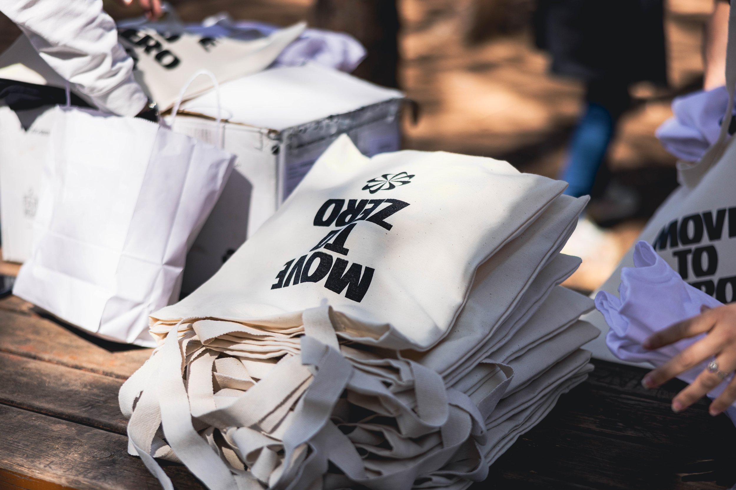 A stack of white tote bags with black text, placed on a wooden surface outdoors, with other items and people partially visible in the background.