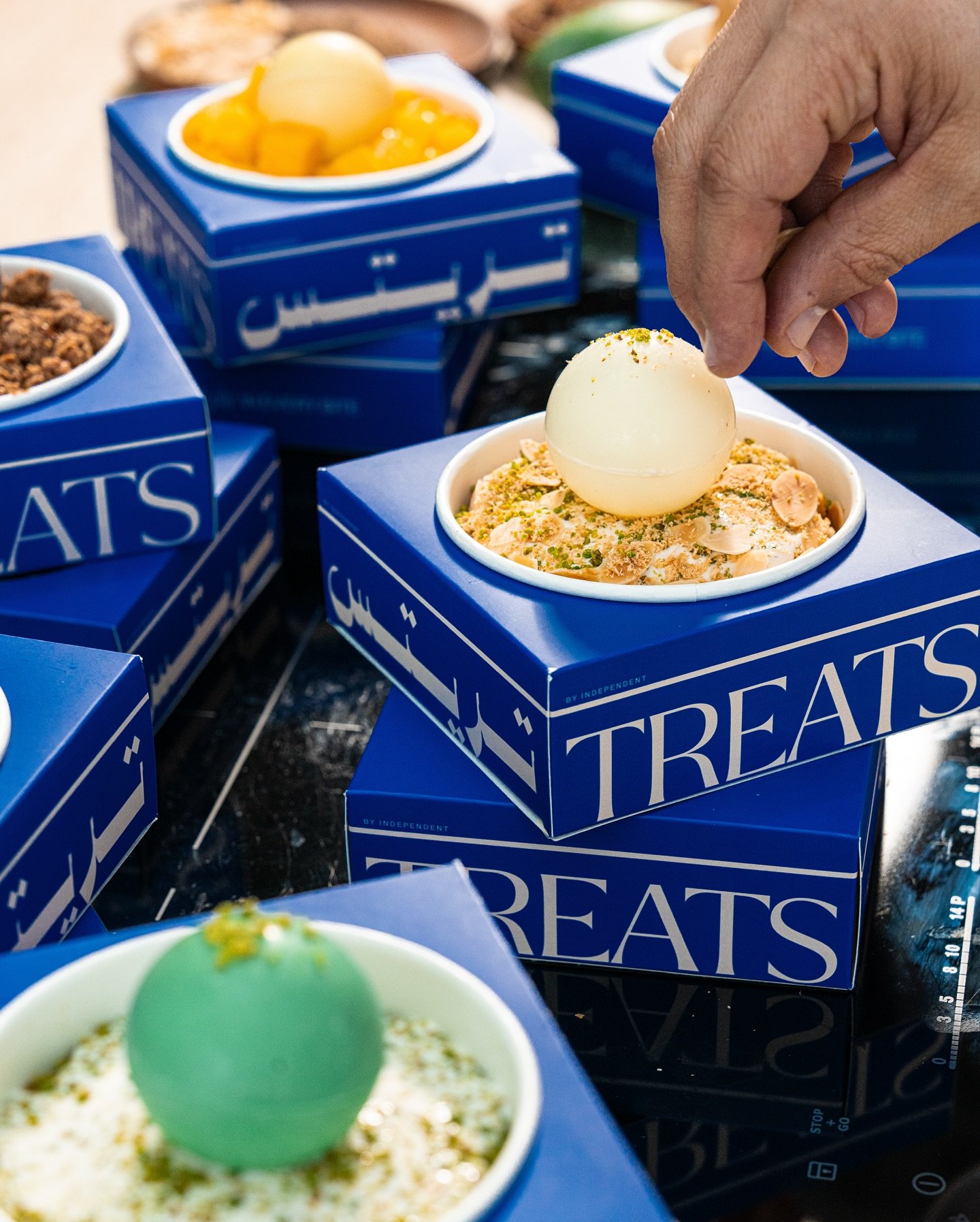 A person decorates ice cream sundaes with a white chocolate egg on top, in blue-tinted boxes labeled 'Treats'.