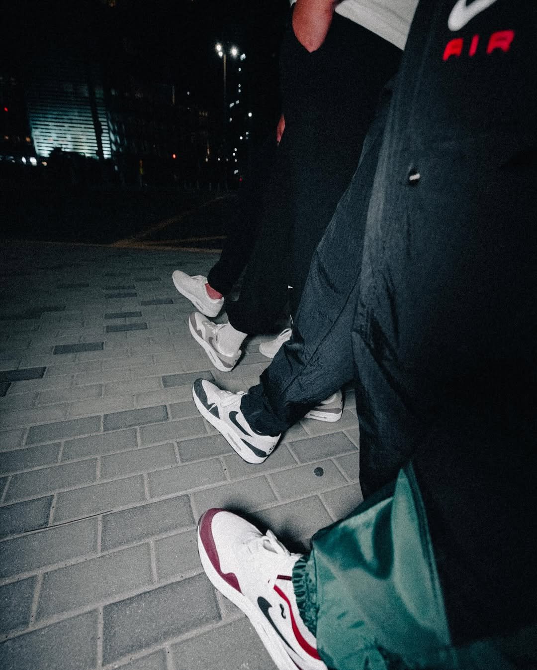 Close-up of people standing on a brick sidewalk at night, showing sneakers and part of their clothing, with city lights in the background.