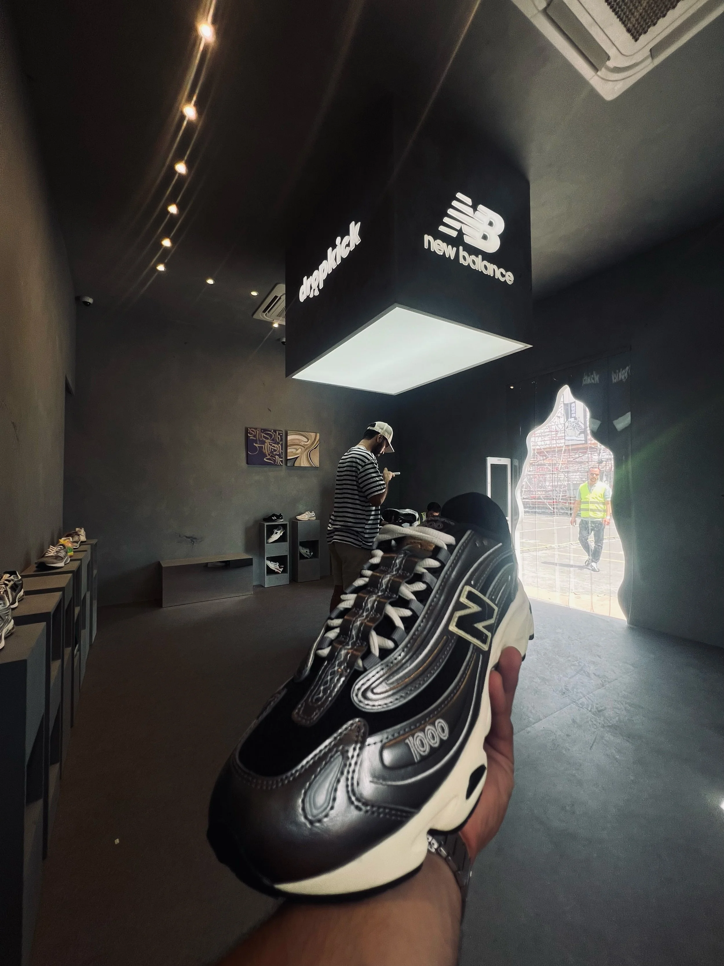 Close-up of a black and white New Balance sneaker held in a person's hand, with a shoe store interior and a person in a striped shirt in the background.