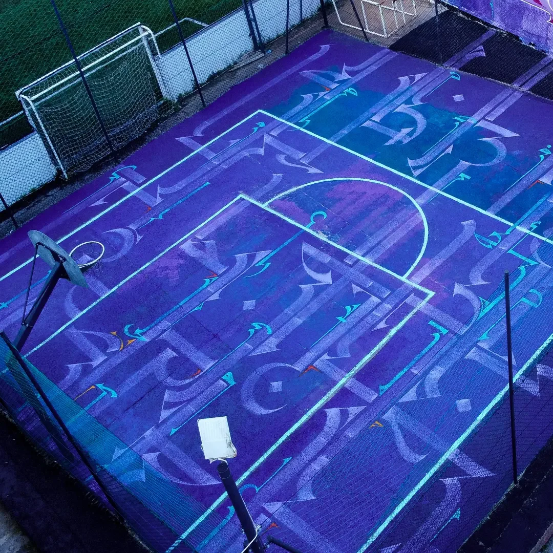 An illuminated basketball court featuring glowing blue and purple lines and artistic geometric patterns on the surface, surrounded by a fence with small soccer goals in the corners.