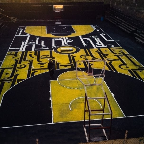 A basketball court with a mural-like design in black, yellow, and white colors, featuring a basketball goal and court markings.