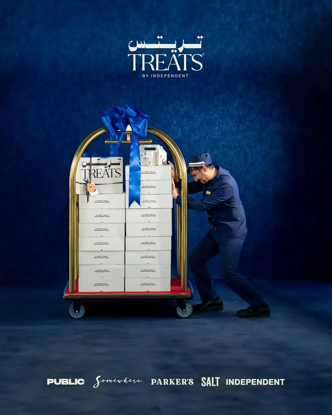 A hotel bellhop in a blue uniform is pushing a luggage cart filled with white boxes labeled 'Treats', with a blue ribbon on top, against a dark blue backdrop with the word 'Treats' in Arabic and English, and logos of PUBLIC, somewhere, PARKER'S SALT, and INDEPENDENT.