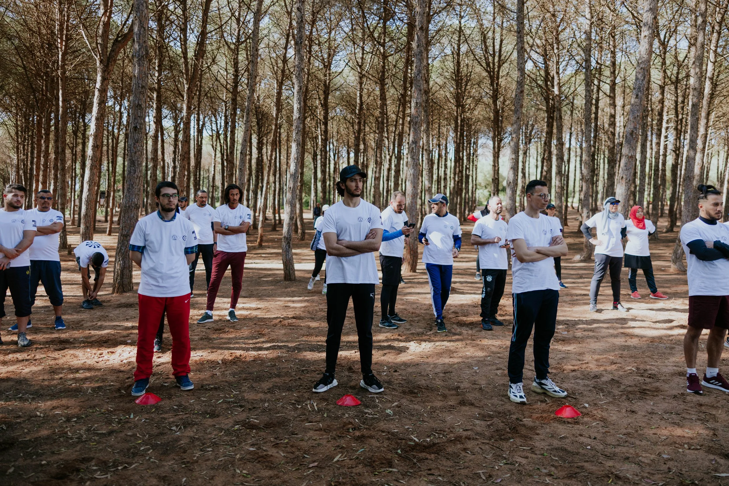 Group of people participating in an outdoor activity in a forest, standing behind red cones arranged on the ground, dressed in casual sportswear.