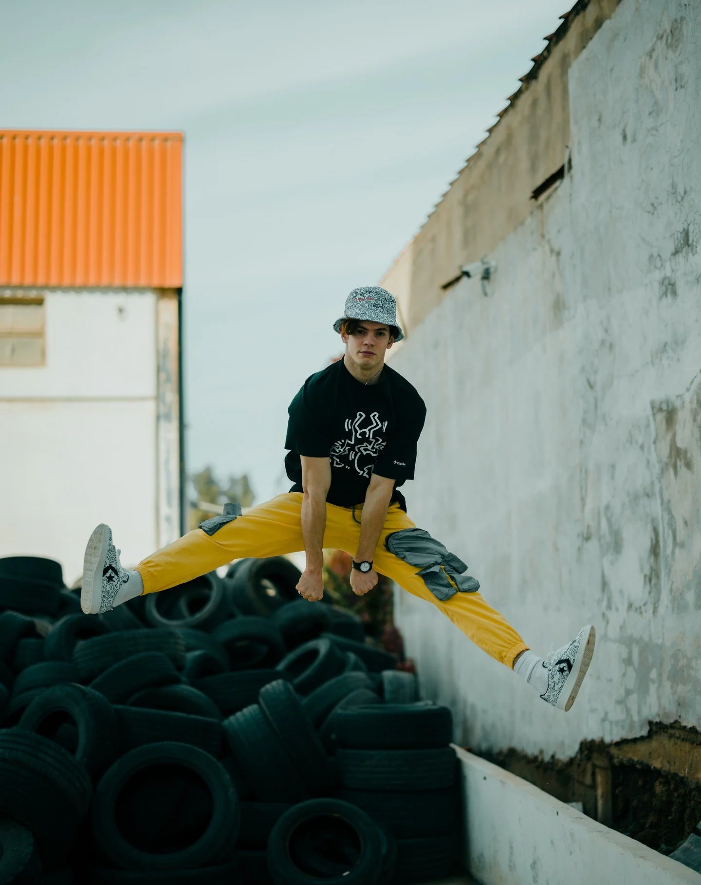 Young man performing a split jump on a pile of tires outdoors, wearing a bucket hat, black t-shirt, yellow pants, and sneakers, next to a weathered wall.