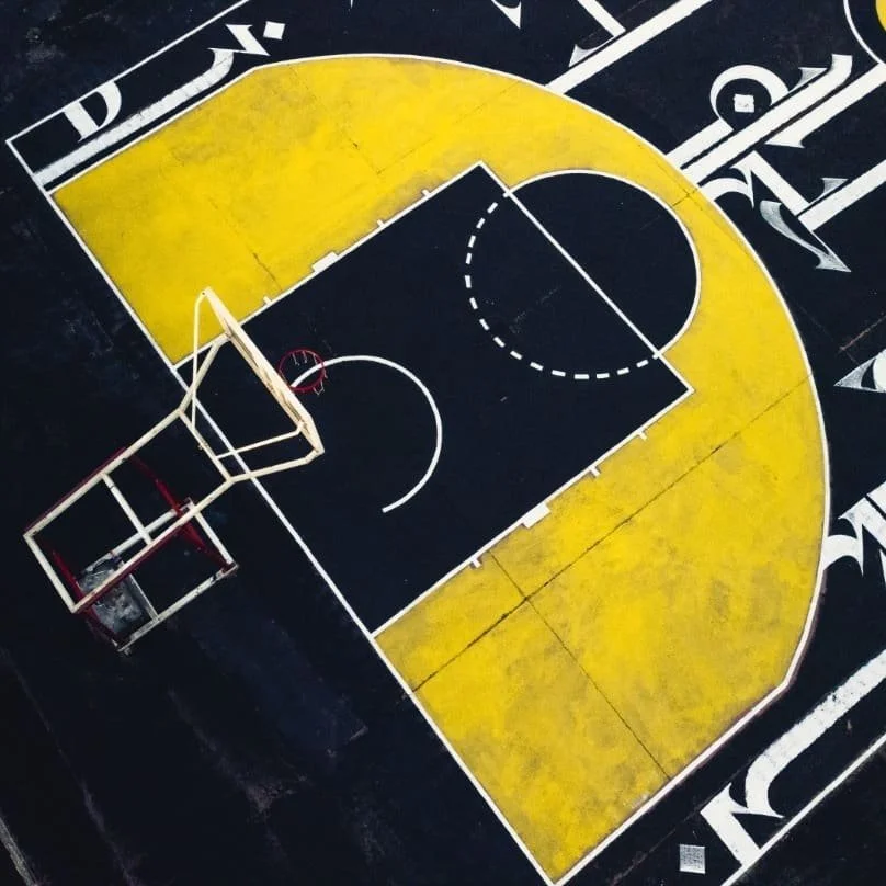An overhead view of a basketball court with yellow and black markings, a hoop with a backboard, and a red and white goal post at one end.