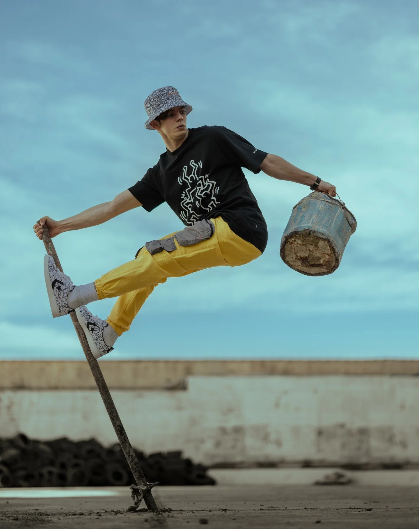 Person balancing on a walking cane while holding a metal bucket, outdoors against a cloudy sky.
