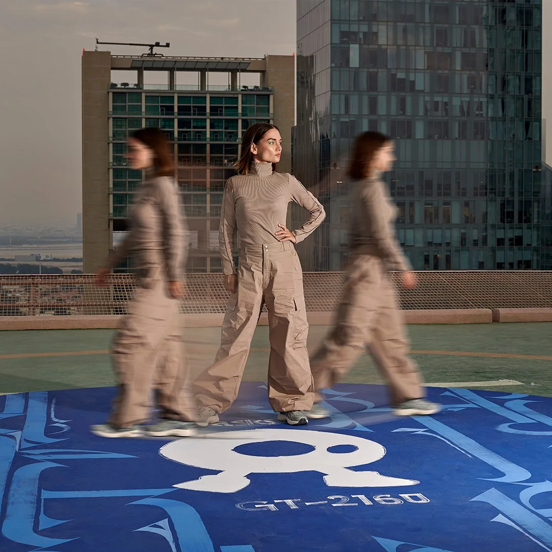 A woman standing on a rooftop with a cityscape background, flanked by two blurred women walking past her, with a blue Nike DC 8-16 logo on the ground.